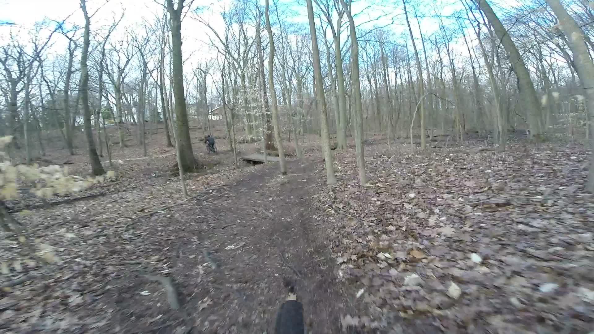 A dirt trail winding through a wooded area with bare trees and scattered fallen leaves. In the background, a person can be seen near a small wooden bridge. The sky is partly cloudy, indicating a cool, overcast day. Richmond Avenue and Forest Hill road mountain bike trail.