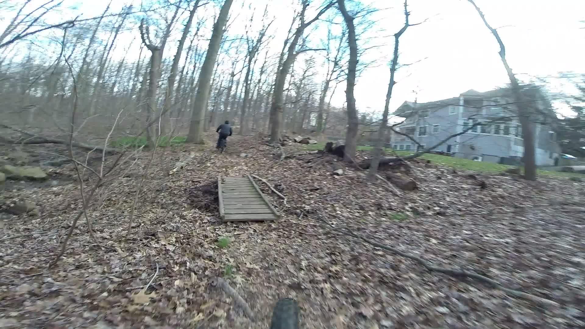 A person riding a bicycle on a forest trail covered in fallen leaves, with trees on either side and a house visible in the background. A wooden bridge crosses a small area along the path. Richmond Avenue and Forest Hill road mountain bike trail.
