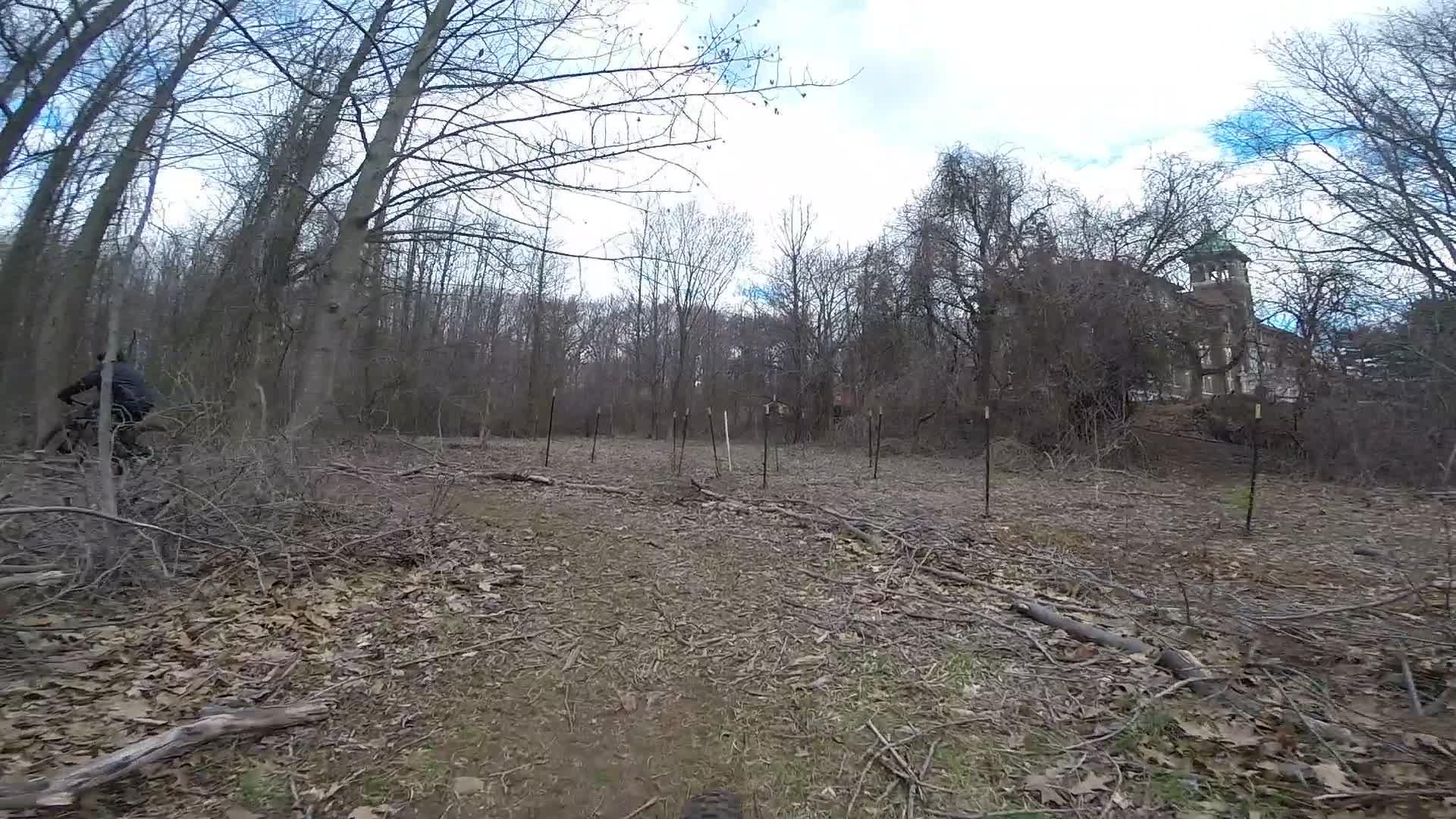 A cyclist riding along a narrow dirt path in a wooded area, surrounded by bare trees and fallen branches, with a distant structure partially obscured by foliage in the background. The sky is overcast, suggesting a cool, early spring day. Trails seperated by streets mountain bike trail.