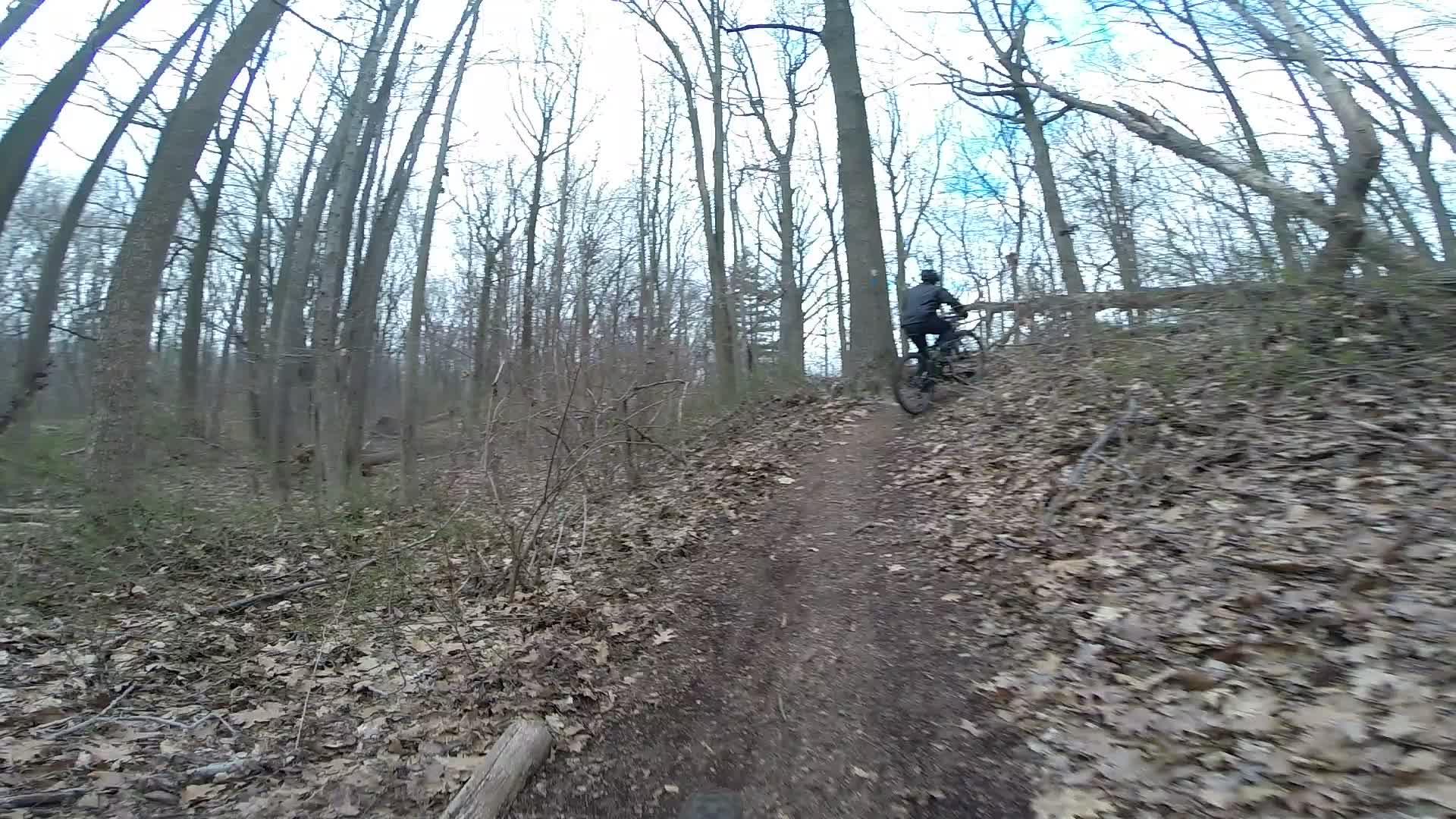 A mountain biker riding up a dirt path surrounded by trees in a wooded area during early spring, with brown leaves on the ground. Trails seperated by streets mountain bike trail.