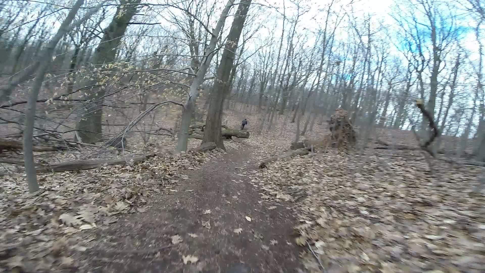 A winding dirt trail through a wooded area, surrounded by bare trees with scattered leaves on the ground. In the background, a person can be seen walking along the path. The sky is partially cloudy. Trails seperated by streets mountain bike trail.