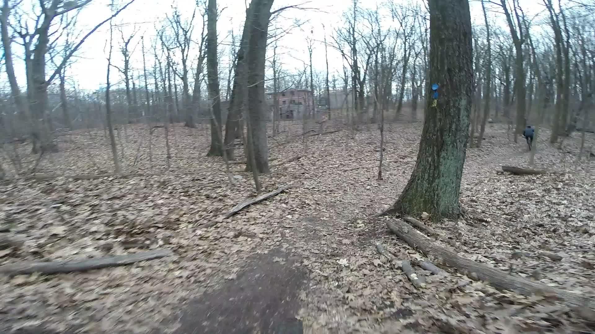 A wooded trail covered with fallen leaves, winding through a forest of bare trees. In the background, a rustic building is partially visible among the trees. A person on a bicycle can be seen riding along the path. The scene is overcast, giving it a muted and tranquil atmosphere. Trails seperated by streets mountain bike trail.