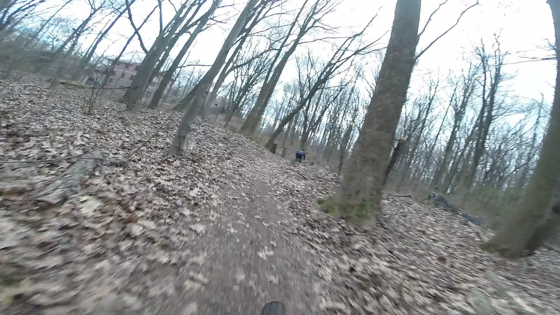 A view from a cyclist's perspective on a dirt trail winding through a forest. The ground is covered with fallen leaves, and trees with bare branches are visible on either side. In the background, a person can be seen biking along the trail. The scene conveys a sense of outdoor adventure in a natural setting. Trails seperated by streets mountain bike trail.