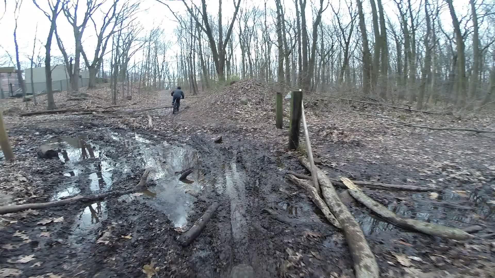 A cyclist navigating a muddy, leaf-covered trail in a wooded area. The path is marked by puddles and scattered logs, with trees in the background indicating an early spring or late winter setting. Trails seperated by streets mountain bike trail.