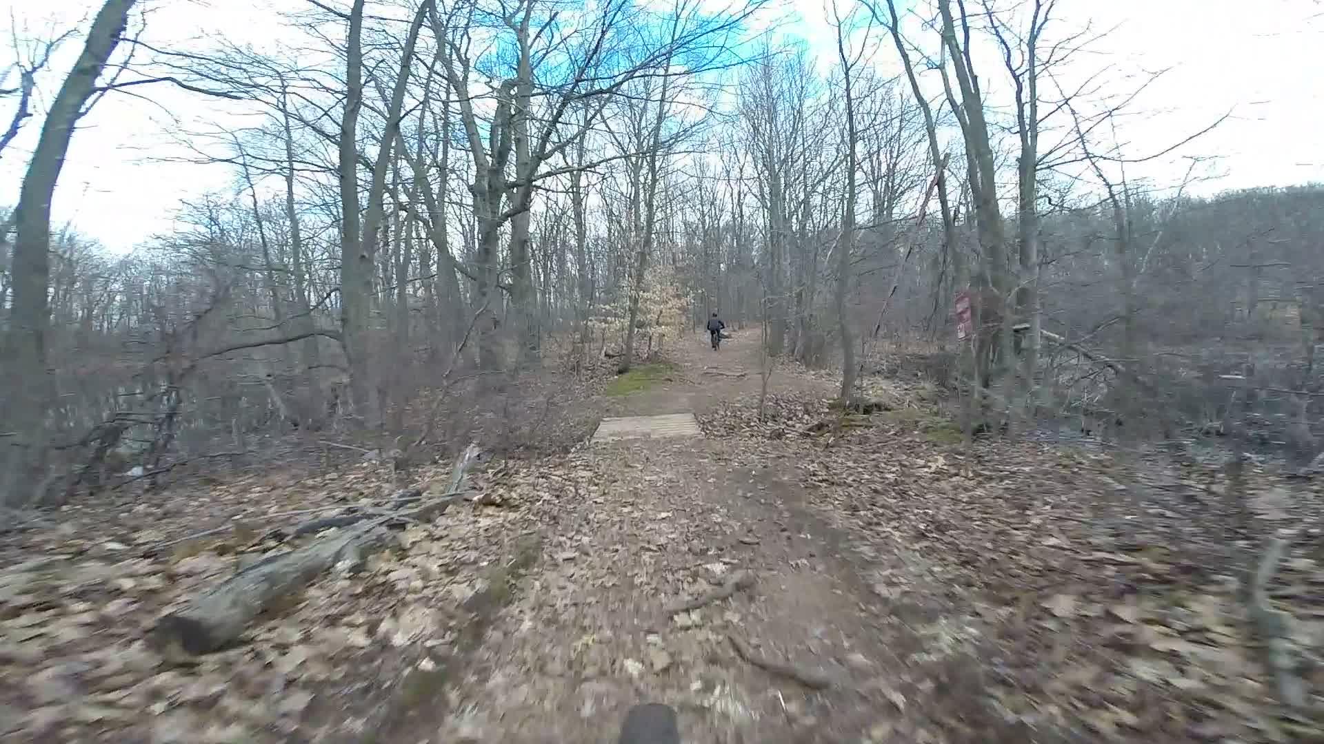 A winding dirt path through a wooded area, with bare trees and scattered fallen leaves on the ground. A cyclist can be seen riding along the trail in the distance, and a wooden bridge crosses a small section of the path. The sky is partly cloudy, suggesting early spring or late autumn conditions. Trails seperated by streets mountain bike trail.