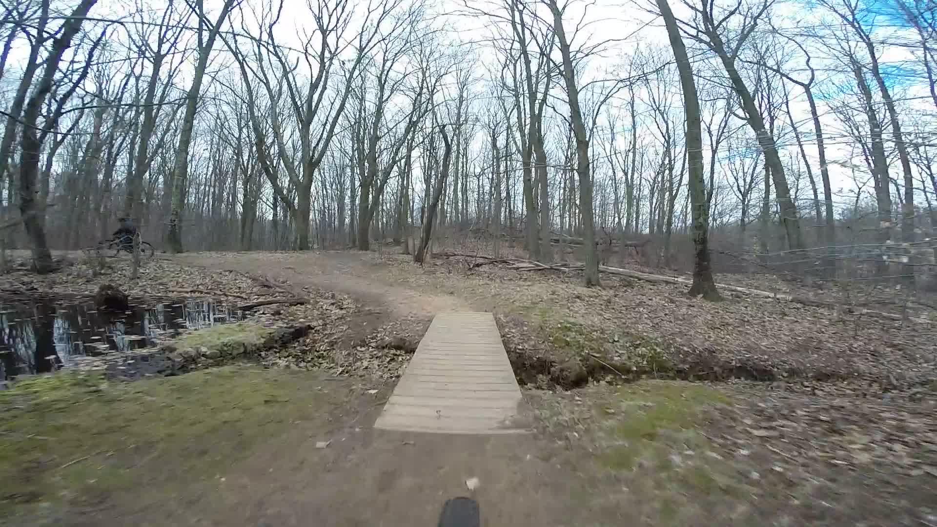 A narrow wooden bridge crosses a small ditch in a wooded area. The scene features bare trees and a path surrounded by fallen leaves, indicating an early spring or late winter setting. A glimpse of water can be seen near the bridge, reflecting the surrounding nature. Trails seperated by streets mountain bike trail.