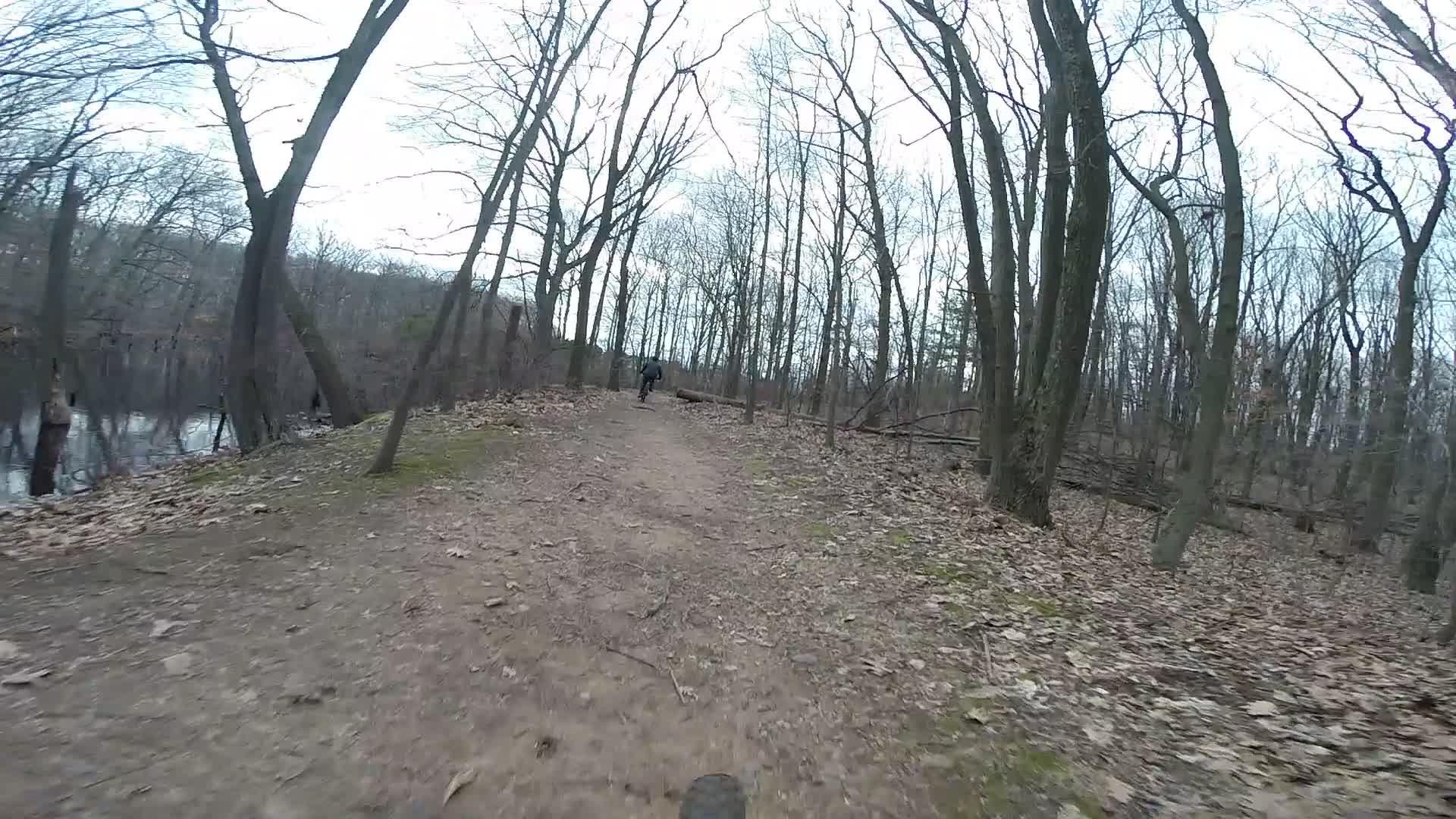 A dirt path winding through a wooded area with bare trees and patches of moss, alongside a calm body of water. A person on a bicycle is visible in the distance, riding along the trail. The scene suggests a mild, overcast day, with fallen leaves scattered on the ground. Trails seperated by streets mountain bike trail.