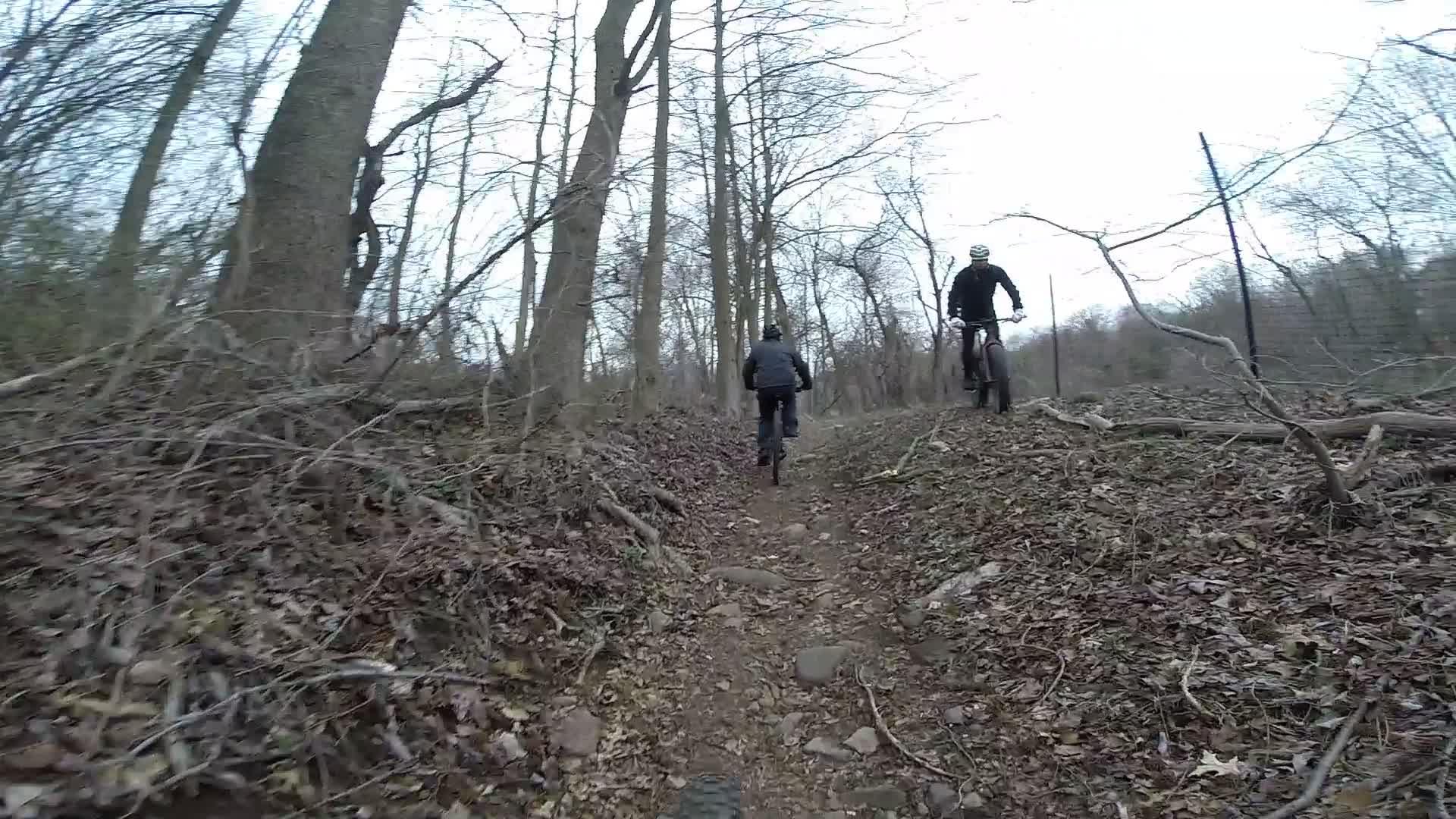 Two cyclists navigate a rocky, leaf-covered trail through a wooded area in early spring. One cyclist is slightly ahead, wearing a helmet and dark jacket, while the other follows behind on a mountain bike. The trees in the background are bare, indicating a cool, overcast day. Richmond Avenue and Forest Hill road mountain bike trail.