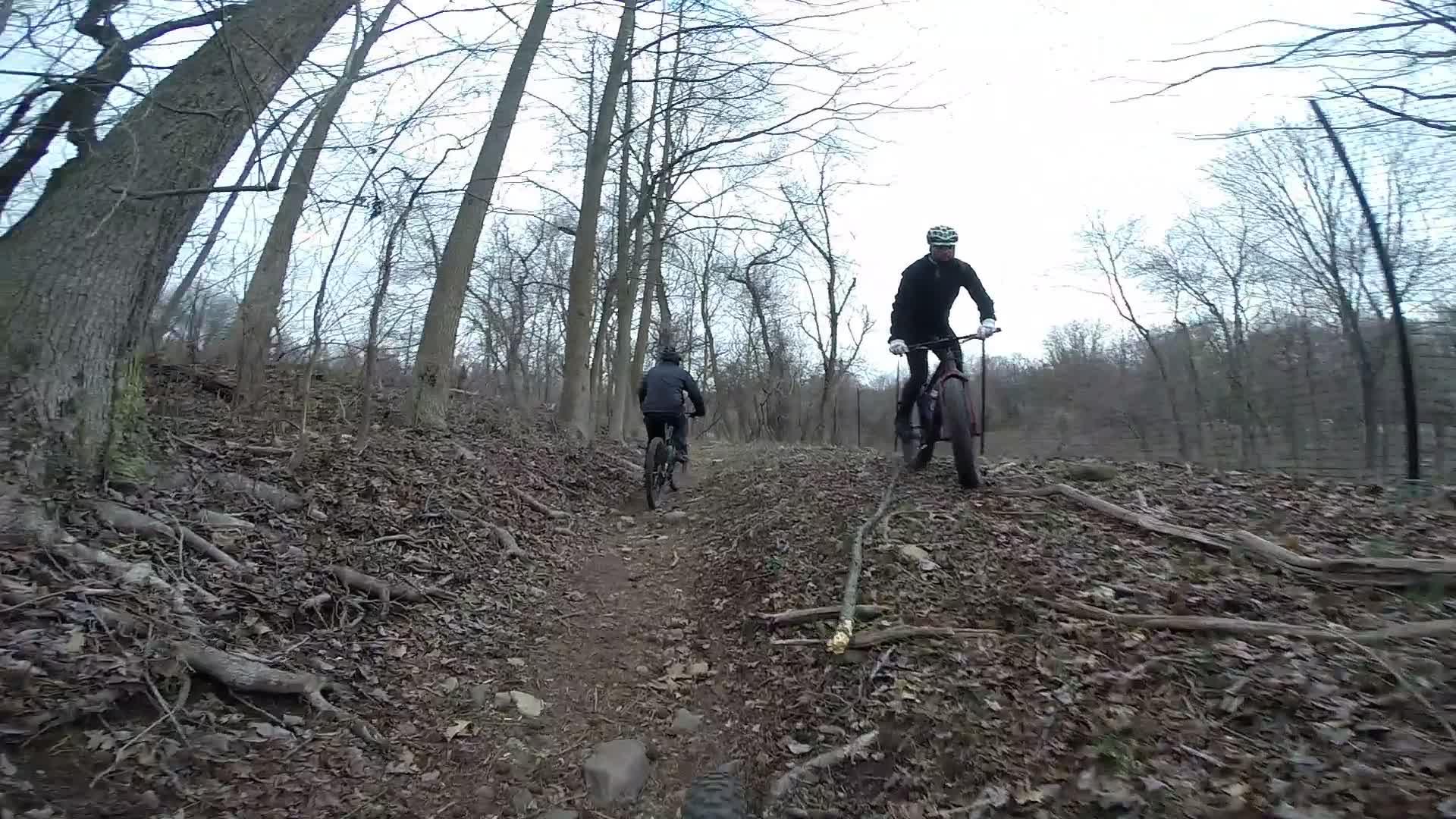 Two mountain bikers ride along a wooded trail in a forested area. The path is lined with leaves, rocks, and tree roots, while bare trees loom overhead against a cloudy sky. The scene captures the essence of outdoor adventure and cycling in nature. Richmond Avenue and Forest Hill road mountain bike trail.