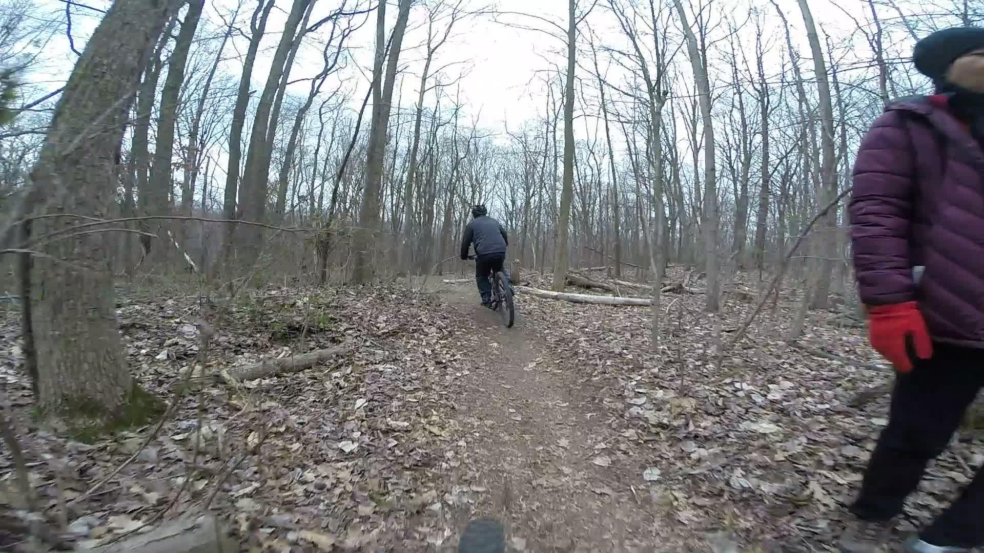 A person riding a mountain bike on a dirt trail through a wooded area during the winter. A second person stands nearby, wearing a purple jacket and red gloves, observing the cyclist. The scene features bare trees and scattered leaves on the ground, indicating a cool, possibly overcast day. Richmond Avenue and Forest Hill road mountain bike trail.