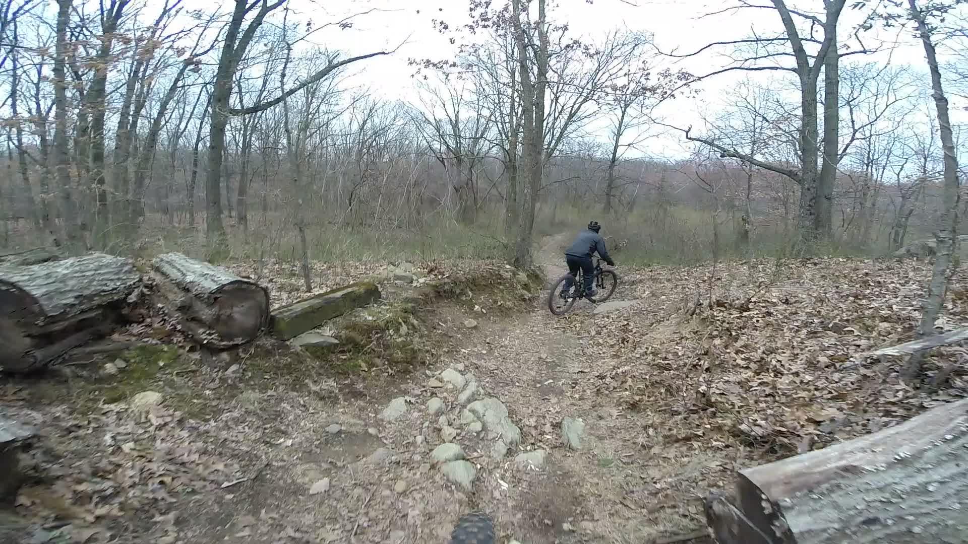 A mountain biker navigating a rocky trail in a wooded area during early spring, with bare trees and fallen leaves visible on the ground. Richmond Avenue and Forest Hill road mountain bike trail.