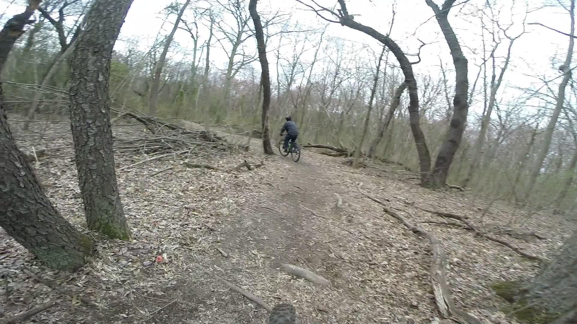 A mountain biker riding on a narrow dirt trail surrounded by bare trees and fallen branches in a wooded area. The ground is covered with dried leaves, and the atmosphere is calm and tranquil. Richmond Avenue and Forest Hill road mountain bike trail.
