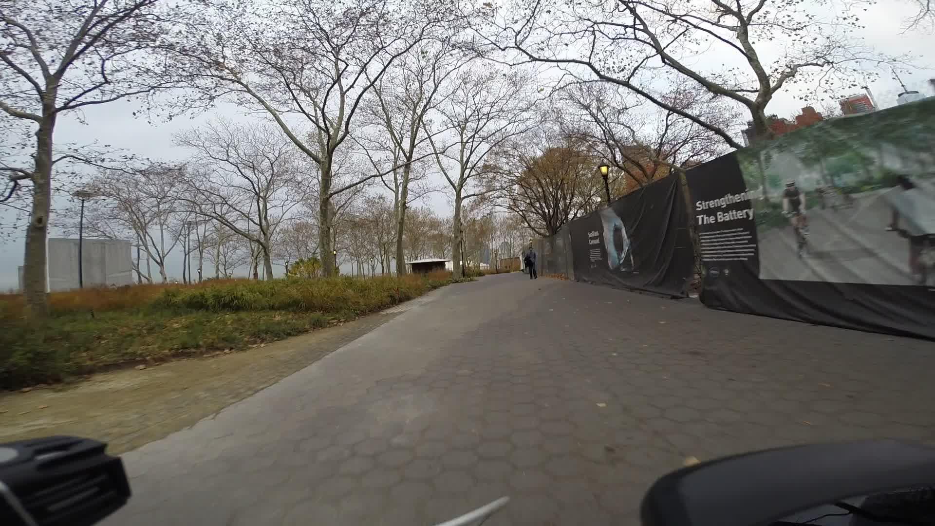 A path lined with bare trees leads through a park, with a construction fence featuring informational signage on one side. The scene captures a cloudy day with muted colors, hinting at autumn. A person is walking along the path, suggesting a quiet atmosphere in a public space. West Street Greenway mountain bike trail.