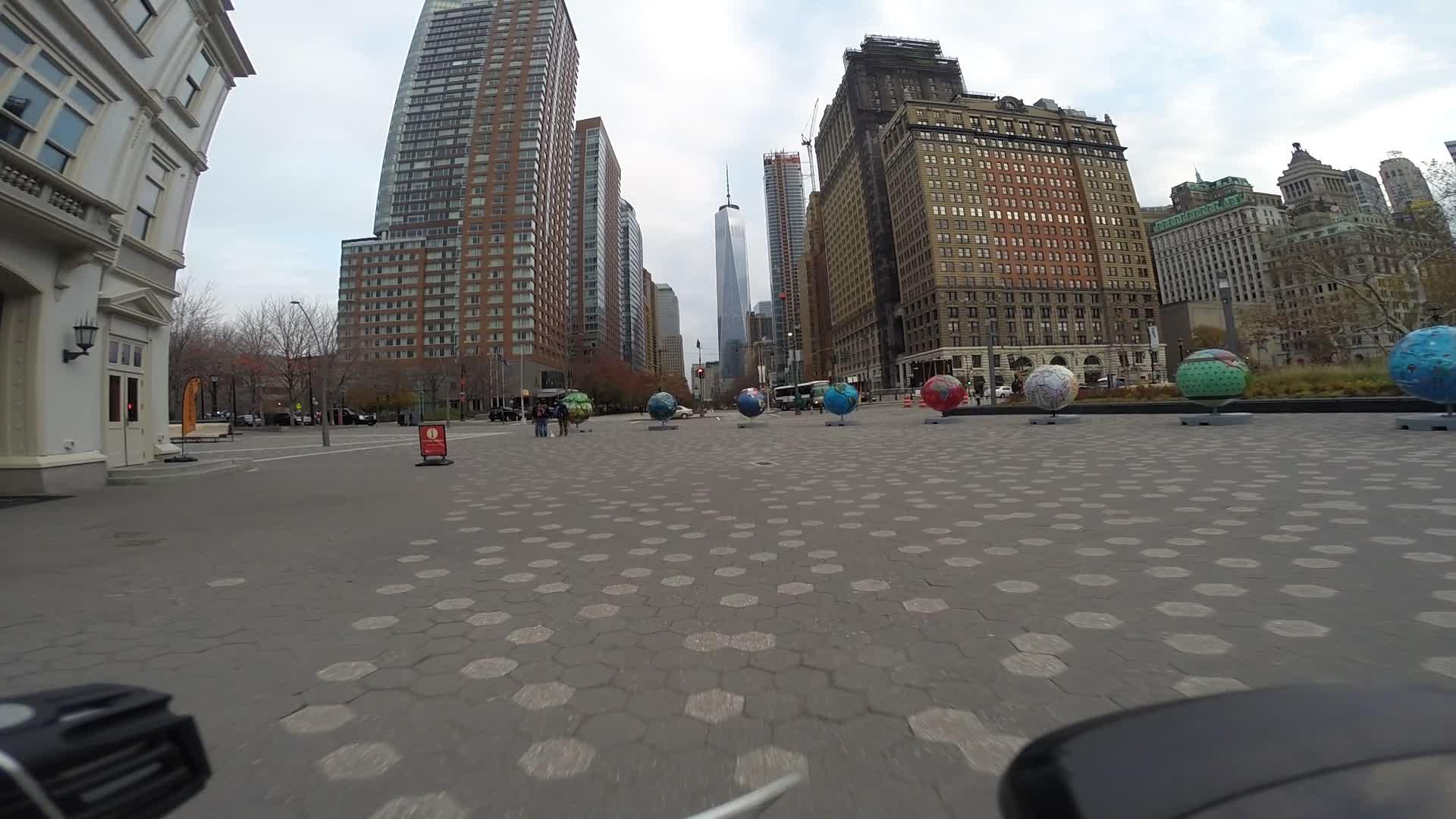 A bustling city street featuring a view of several large, colorful globe sculptures arranged along a pavement with honeycomb patterns. In the background, tall skyscrapers, including a prominent tower, rise against a cloudy sky. Pedestrians can be seen interacting in the area, suggesting a lively urban environment. West Street Greenway mountain bike trail.