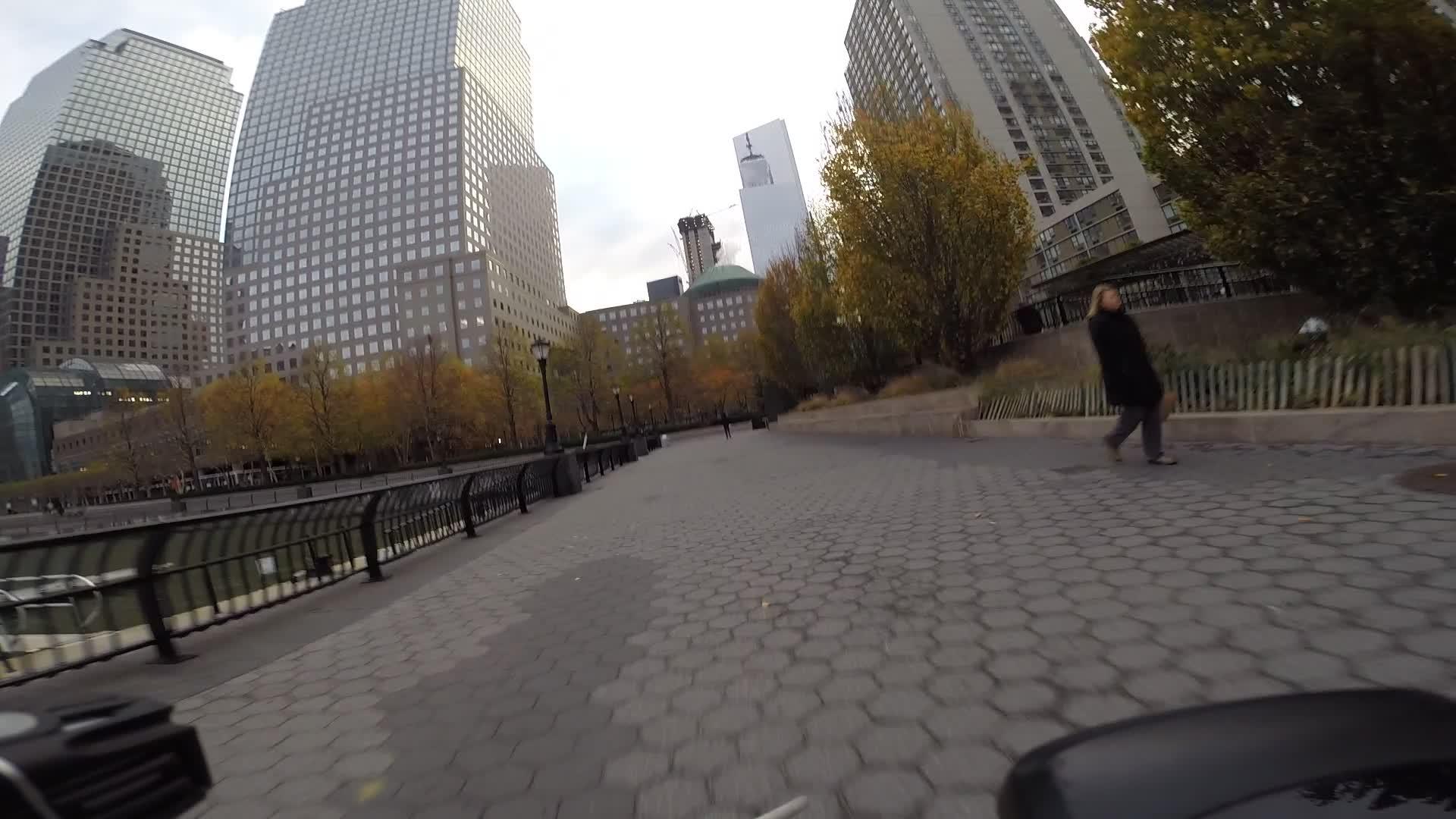 A pedestrian walks along a paved pathway lined with trees displaying autumn foliage, with tall modern buildings reflecting the sky in the background. The scene captures an urban environment near a waterway. West Street Greenway mountain bike trail.