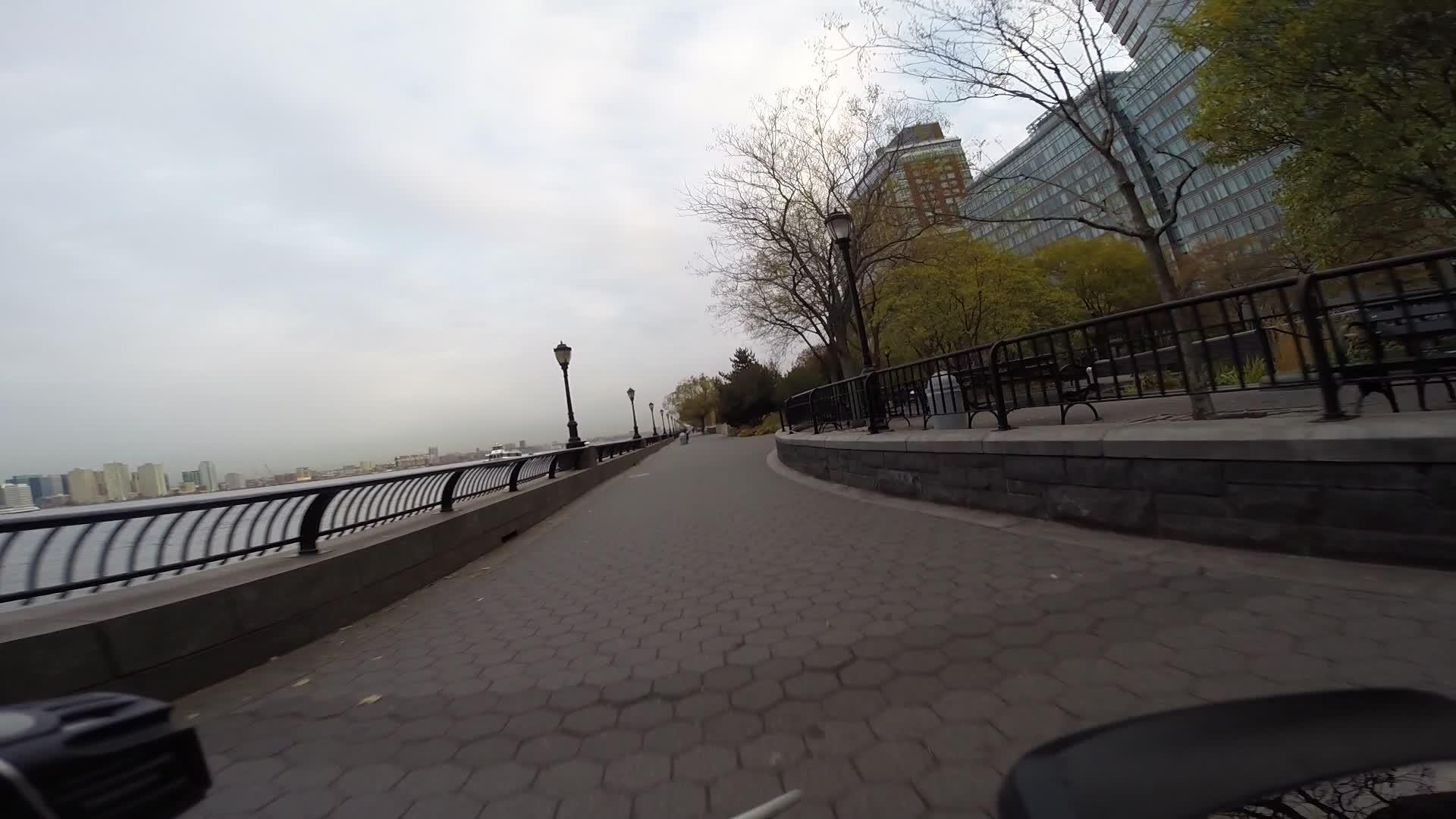 Scenic walkway along a waterfront lined with trees and benches, featuring a path made of hexagonal tiles, with city buildings visible in the background under a cloudy sky. West Street Greenway mountain bike trail.