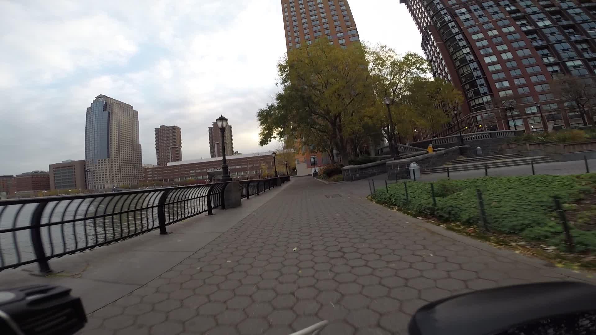 A pathway alongside a river, featuring a brick walkway lined with a black railing. In the background, a skyline of tall buildings is visible, with one glass structure standing out. Trees with autumn foliage and a cloudy sky overhead complete the scene. West Street Greenway mountain bike trail.