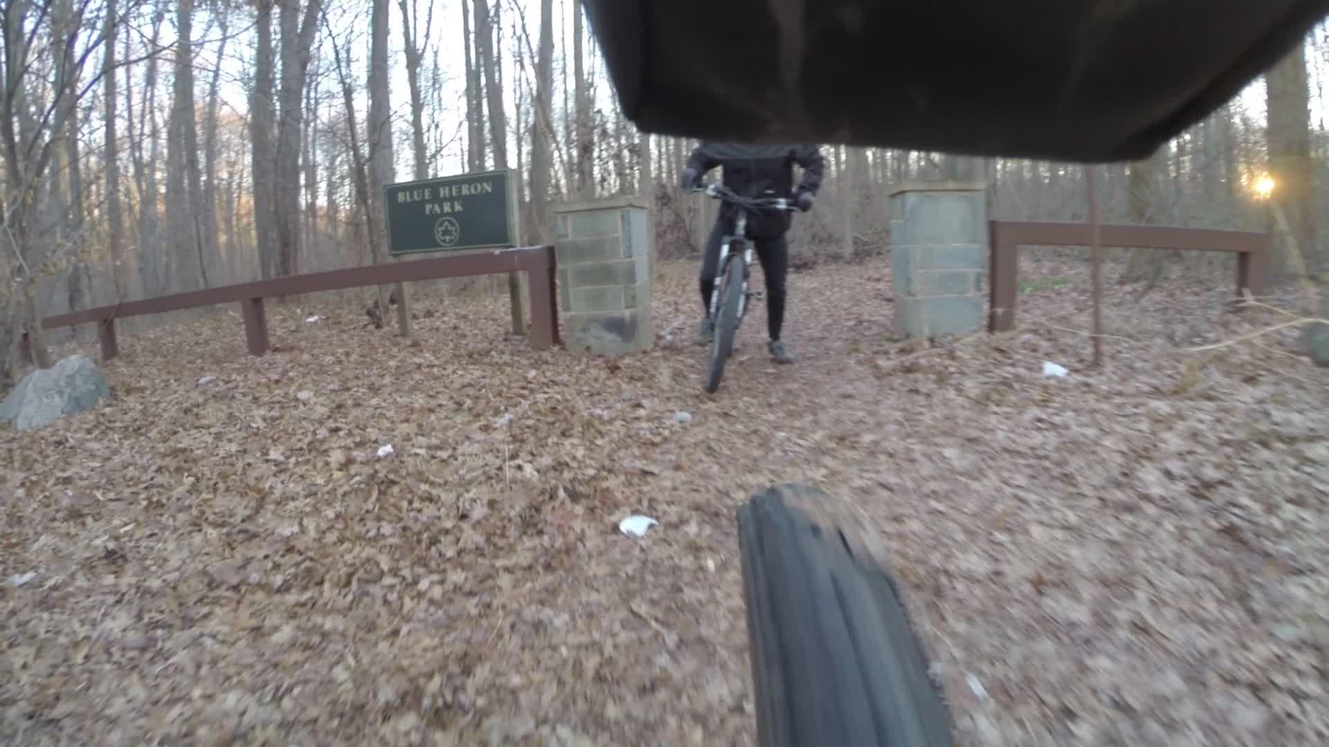 A mountain biker approaches the entrance to Blue Heron Park, surrounded by trees and autumn leaves on the ground. The park sign is visible in the background, indicating the location. Trails seperated by streets mountain bike trail.