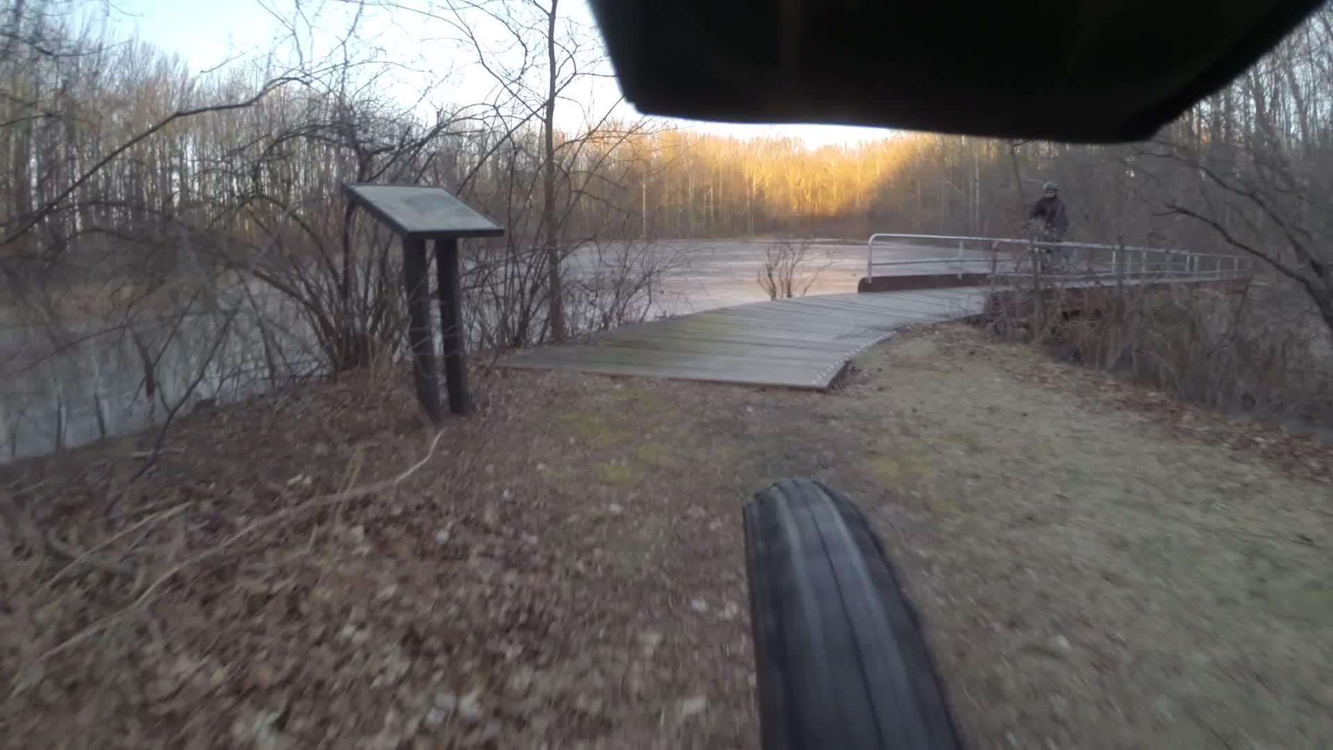 Close-up view of a bicycle tire rolling along a path near a riverbank, with a wooden bridge visible in the background. A sign board is positioned on the left side, surrounded by bare trees and brush. The scene is captured in early morning light, highlighting the tranquility of the landscape. Trails seperated by streets mountain bike trail.