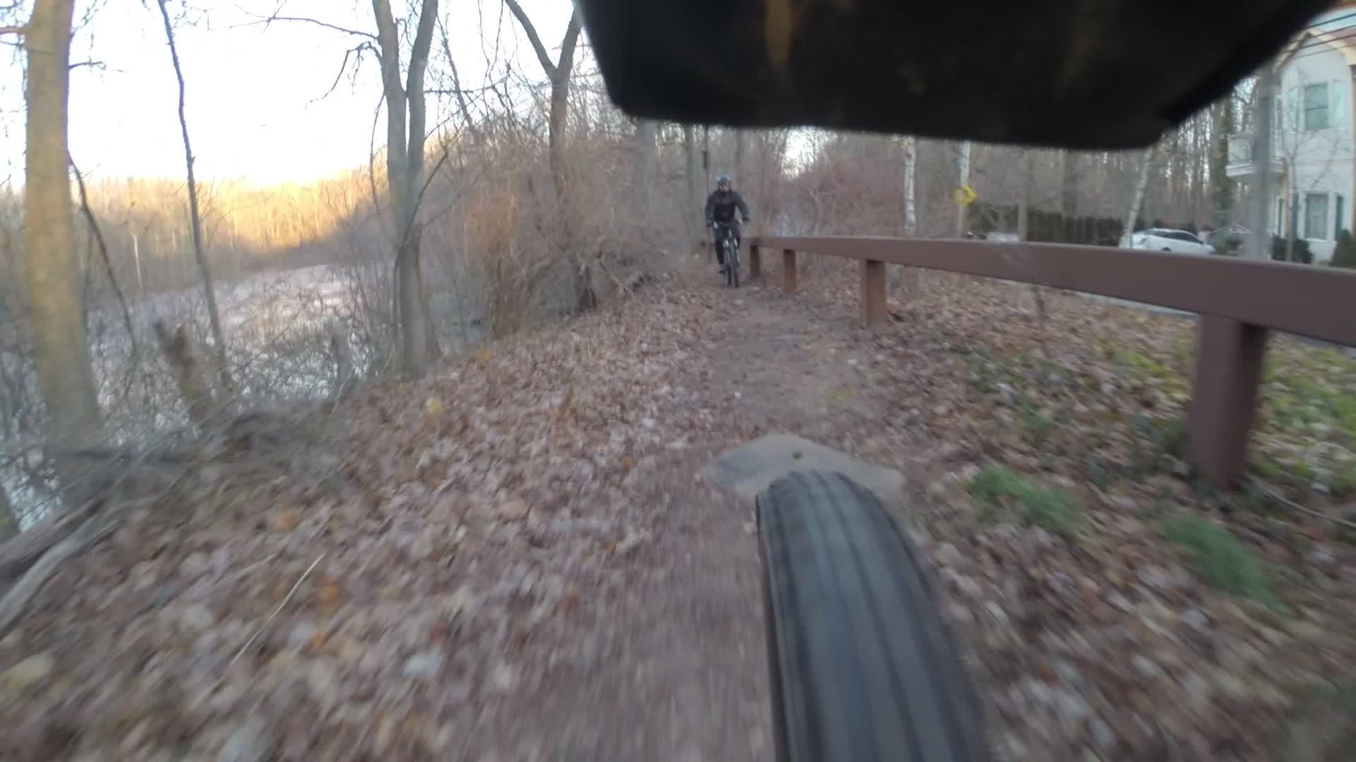 A view from the perspective of a mountain bike tire on a forest trail, with fallen leaves covering the ground. In the background, a cyclist rides along a wooden railing beside a river, surrounded by bare trees and early evening light. Trails seperated by streets mountain bike trail.