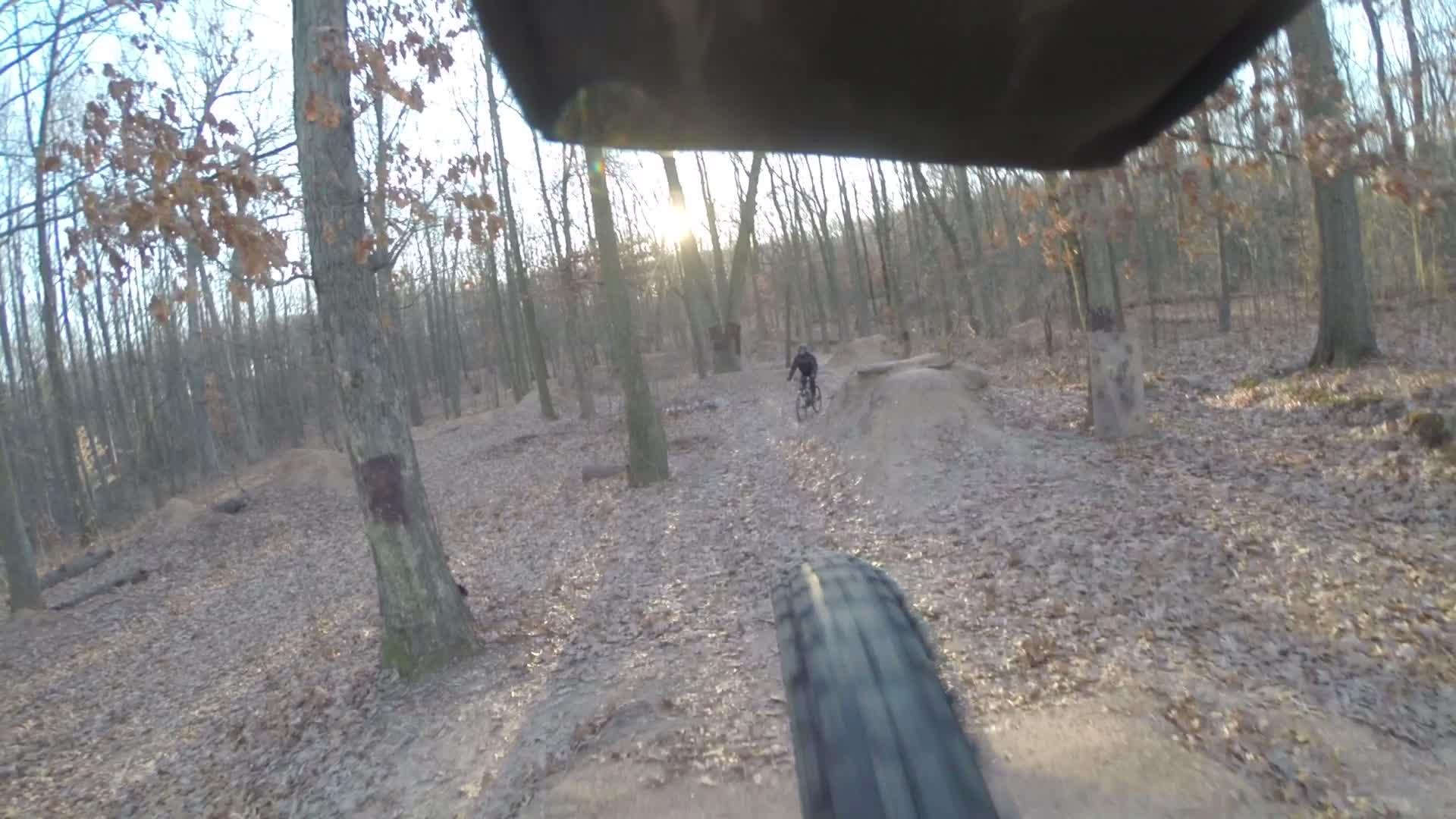 A view from the perspective of a mountain bike tire, showing a dirt trail through a wooded area with trees shedding leaves. A cyclist is visible in the background, approaching a dirt jump as the sun sets, casting a warm light through the trees. Wolfes Pond park mountain bike trail.