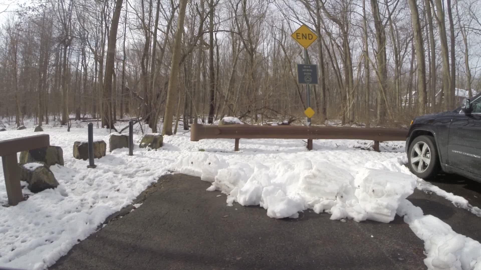 A snowy forest scene depicting a parking area with a "Dead End" sign. The ground is covered in snow, and a brown wooden barrier runs along the edge, next to visible rocks. A black vehicle is parked to the right, surrounded by snow and trees in the background, indicating winter conditions. Trails seperated by streets mountain bike trail.