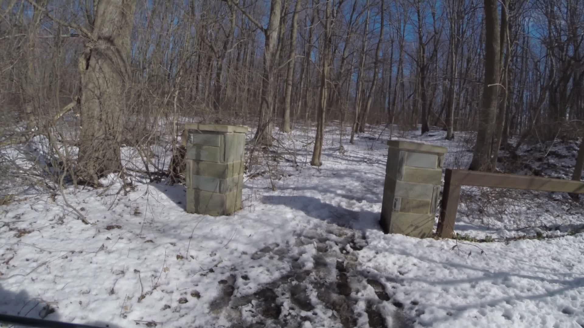 A snowy forest path bordered by two stone pillars, with bare trees in the background and patches of snow on the ground. Sunlight is shining through the branches, creating a serene winter atmosphere. Trails seperated by streets mountain bike trail.