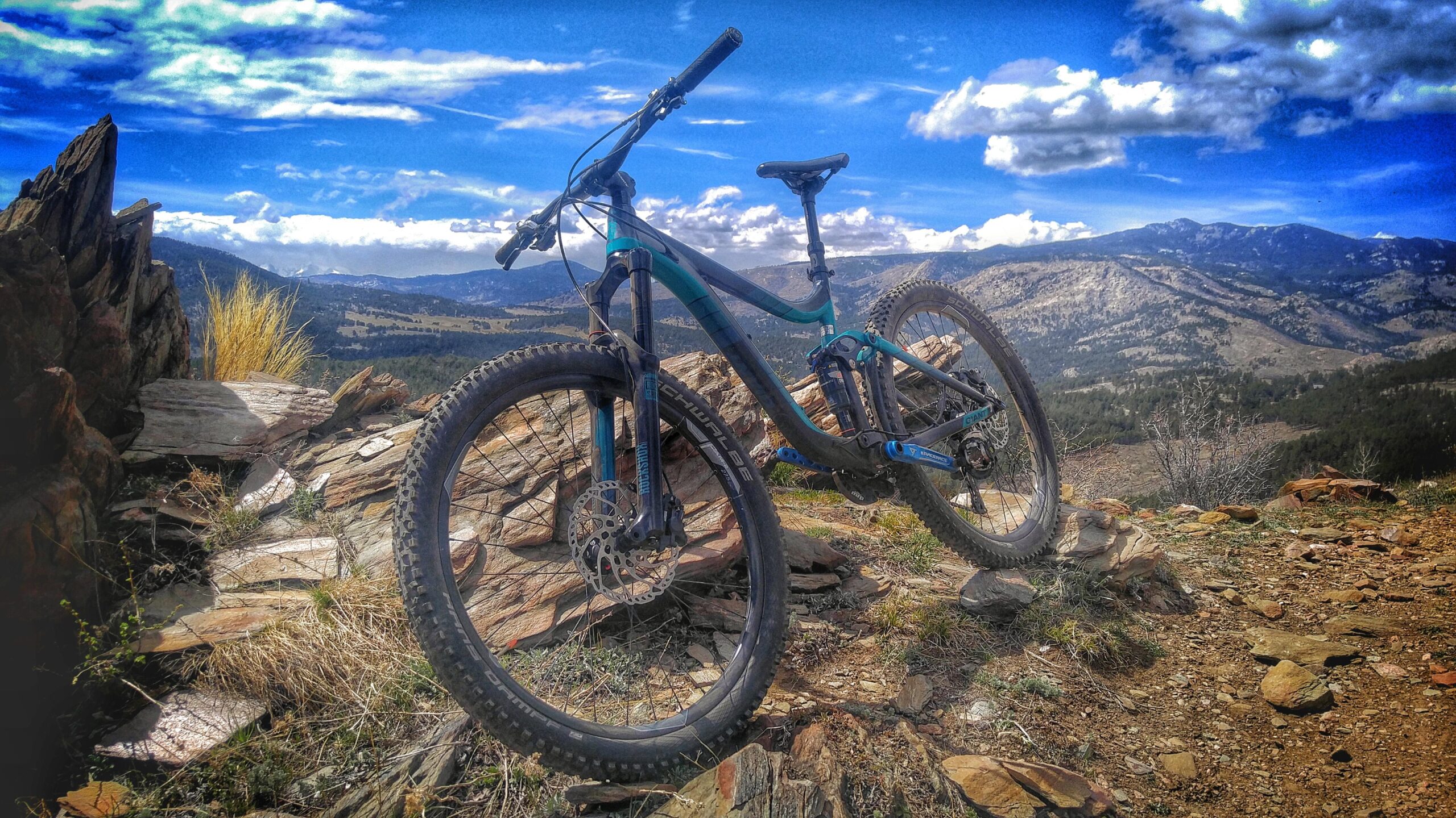 Giant Reign 2: A mountain bike resting on rocky terrain with a scenic view of mountains and blue skies in the background, featuring clouds and patches of grass.