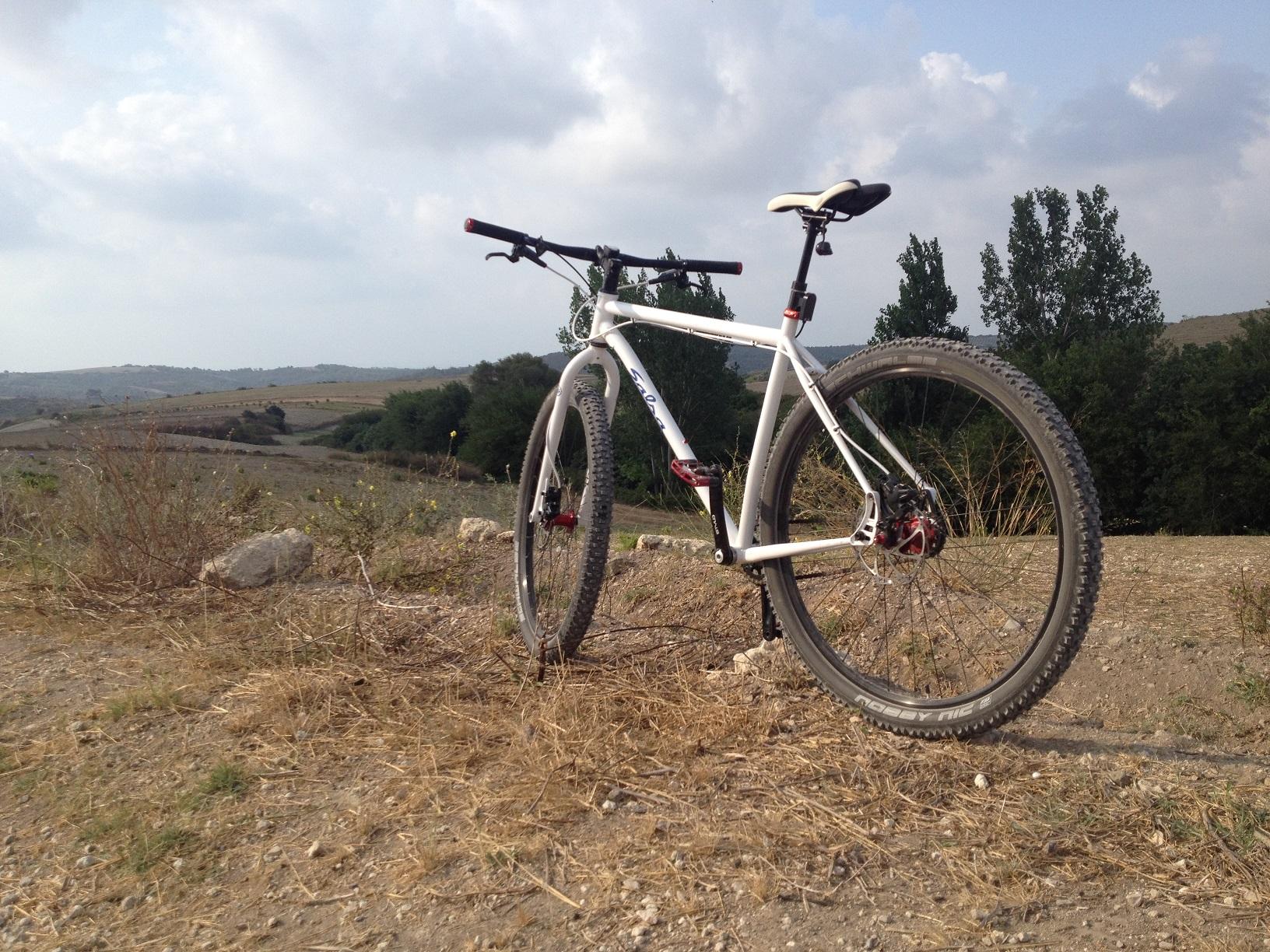 Salsa Salsa El Mariachi SS: A white mountain bike parked on a dirt path surrounded by dry grass and rocky terrain, with rolling hills and trees in the background under a partly cloudy sky.