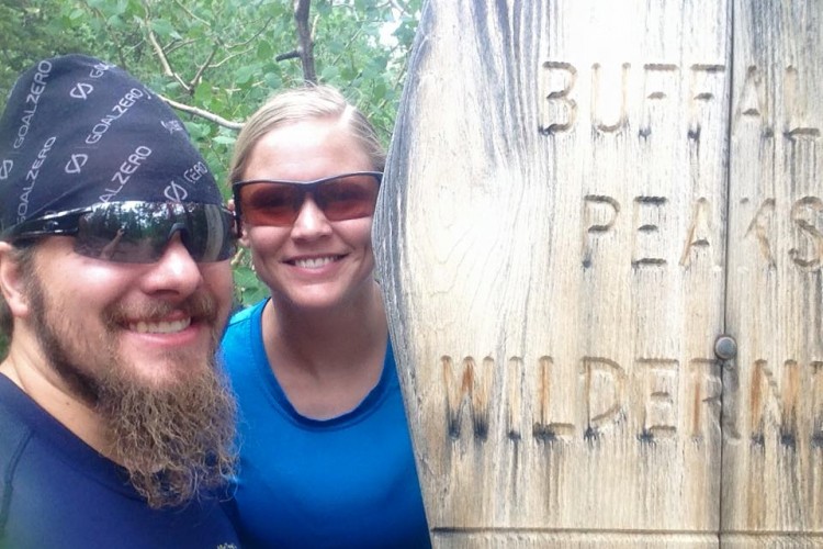 Two people smiling for a selfie in front of a wooden sign that reads "Buffalo Peaks Wilderness." The background features lush greenery typical of a forest. One person is wearing a black bandana and sunglasses, while the other is in a blue shirt and also wearing sunglasses.