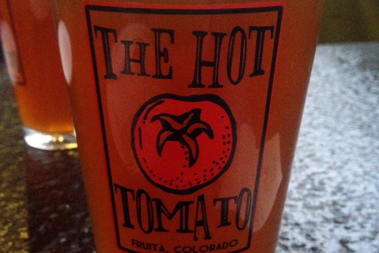 A close-up image of a glass of beer with a logo that reads "THE HOT TOMATO" and "FRUITA, COLORADO." The beer has a golden amber color with a foamy head, sitting on a dark surface. A second glass is faintly visible in the background.