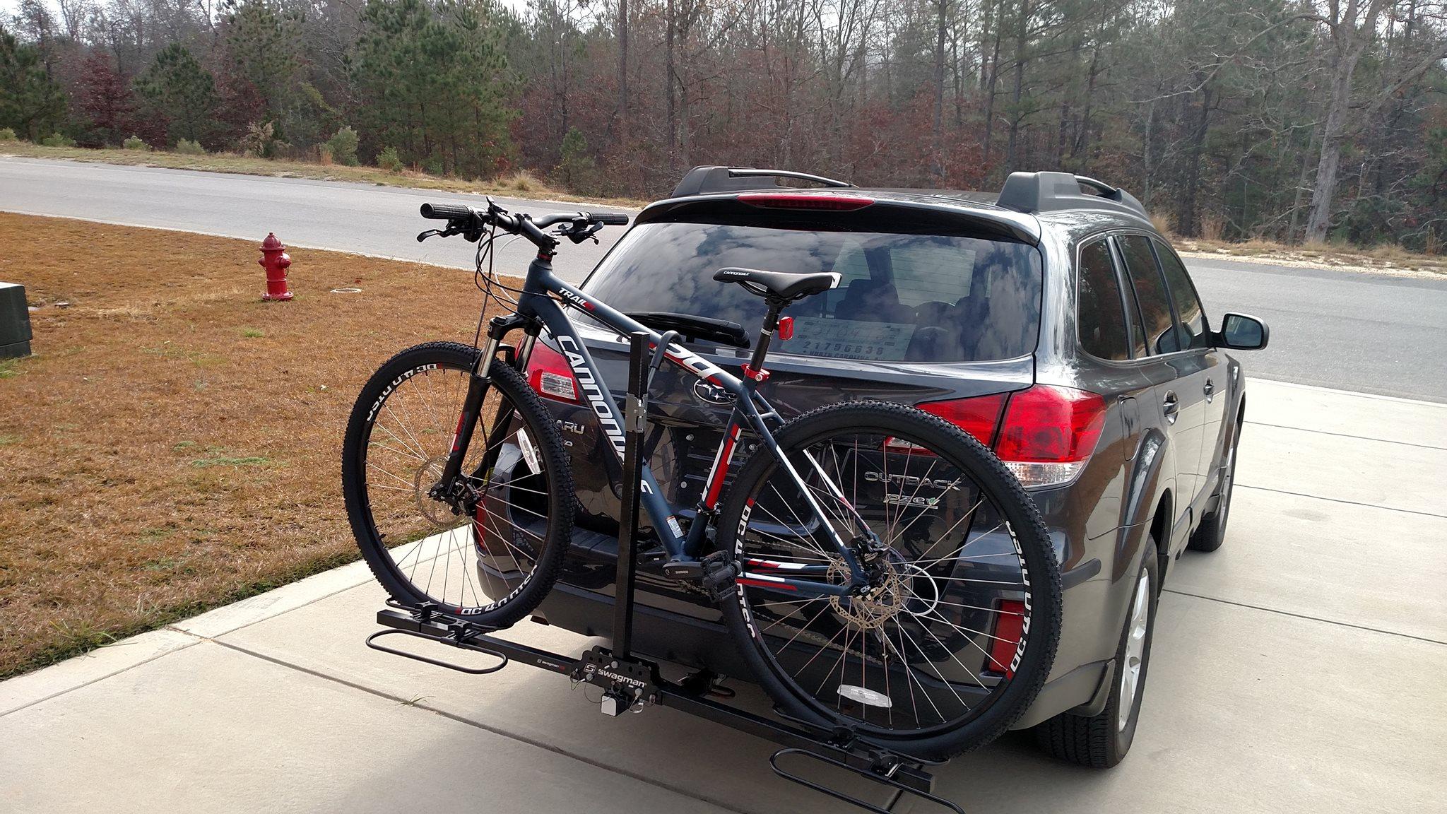 Cannondale Trail 29er 5: A black SUV parked in a driveway with a bike rack mounted on the back, holding two mountain bikes. One bike is prominently displayed, showing its frame and wheels. In the background, there are trees and a small red fire hydrant visible along the road. The driveway is made of concrete and has dry grass around it.