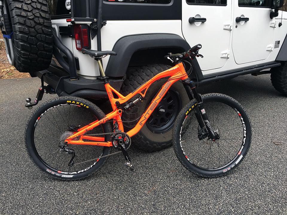 Intense Spider 275: Alt tag: "An orange mountain bike parked next to a black and white off-road vehicle, showcasing the bike's detailed frame and wheels on a paved surface."