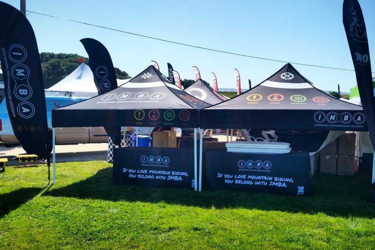 "Outdoor booth for the International Mountain Bicycling Association (IMBA) featuring two black pop-up tents with the IMBA logo and colorful icons representing biking principles. The booth is set up on a green grassy area with banners and promotional materials visible."