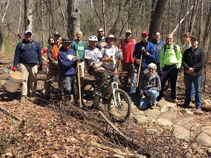 A group of diverse individuals posing together in a wooded area, all wearing outdoor clothing. Some hold tools such as shovels and buckets, while one person is seated on a mountain bike. The scene captures a community effort, possibly related to trail maintenance or environmental conservation, amidst bare trees and fallen leaves on the ground.
