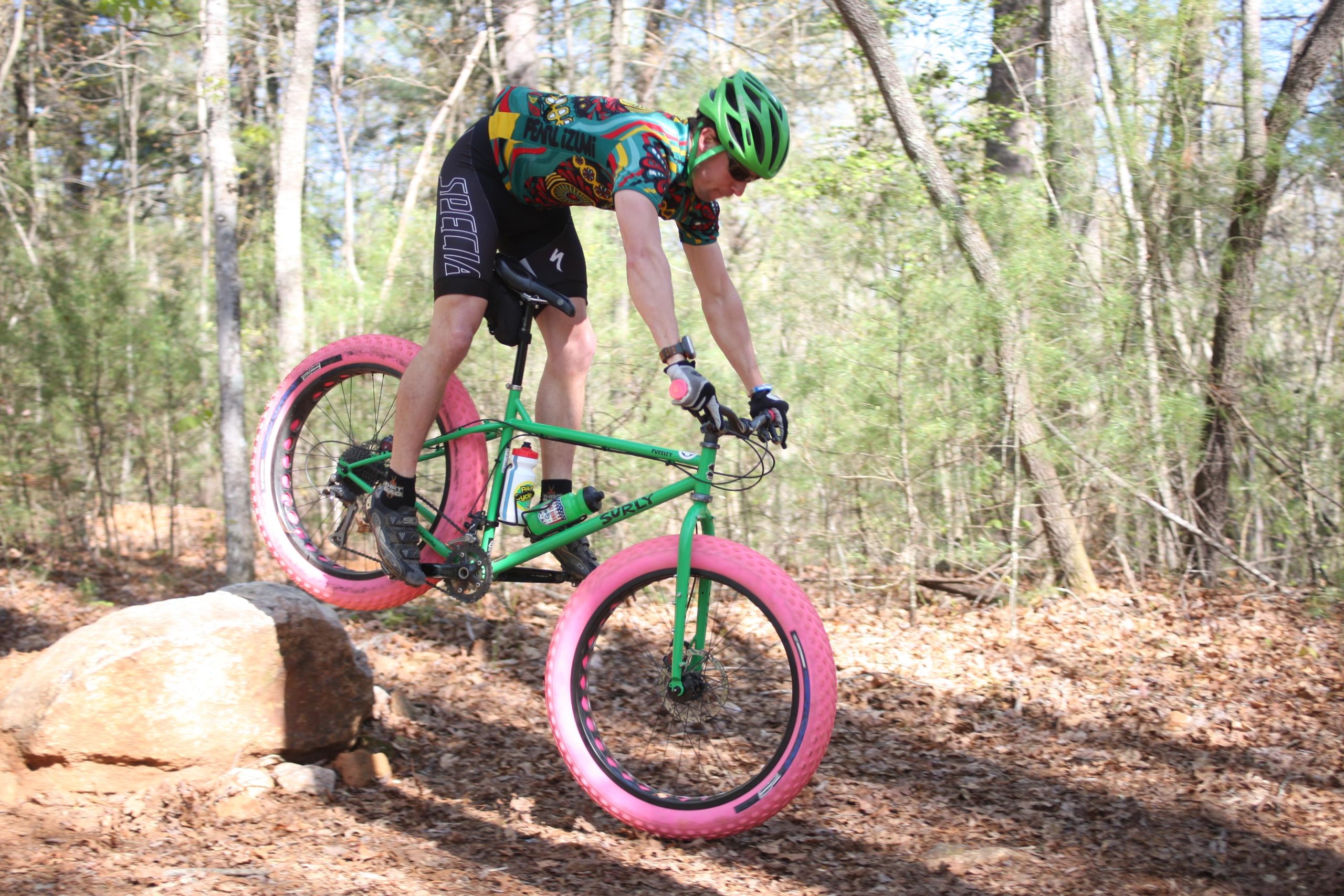 A cyclist wearing a colorful jersey and helmet is performing a jump on a green bike with oversized pink tires while navigating over a rock on a dirt trail surrounded by trees. Dark Mountain Trail mountain bike trail.