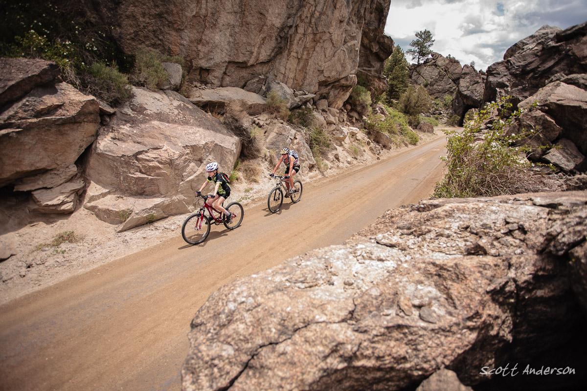 Two cyclists riding mountain bikes on a dirt path surrounded by rocky terrain and vegetation. The scene captures a vibrant outdoor setting under a partly cloudy sky. River Road / #371 mountain bike trail.