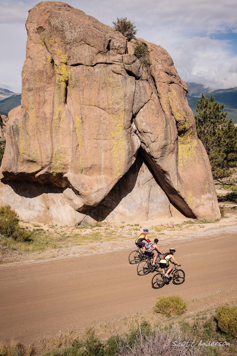 Three cyclists riding along a dirt road near a large rock formation, with green vegetation in the foreground and mountains in the background under a partly cloudy sky. River Road / #371 mountain bike trail.