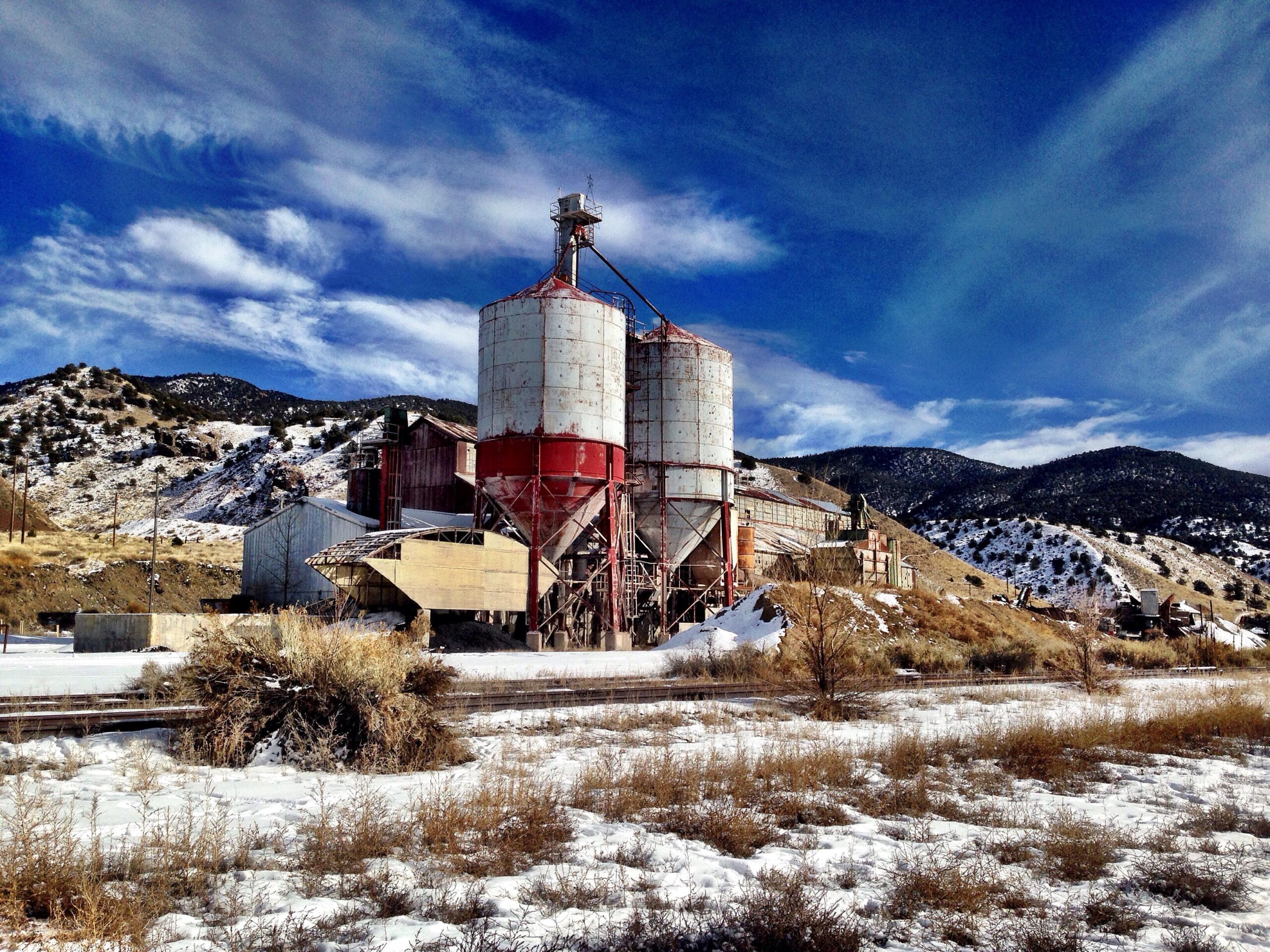 Abandoned industrial facility with red and white silos, surrounded by snow-covered ground and hills in the background under a blue sky with wispy clouds. Arkansas Hills mountain bike trail.