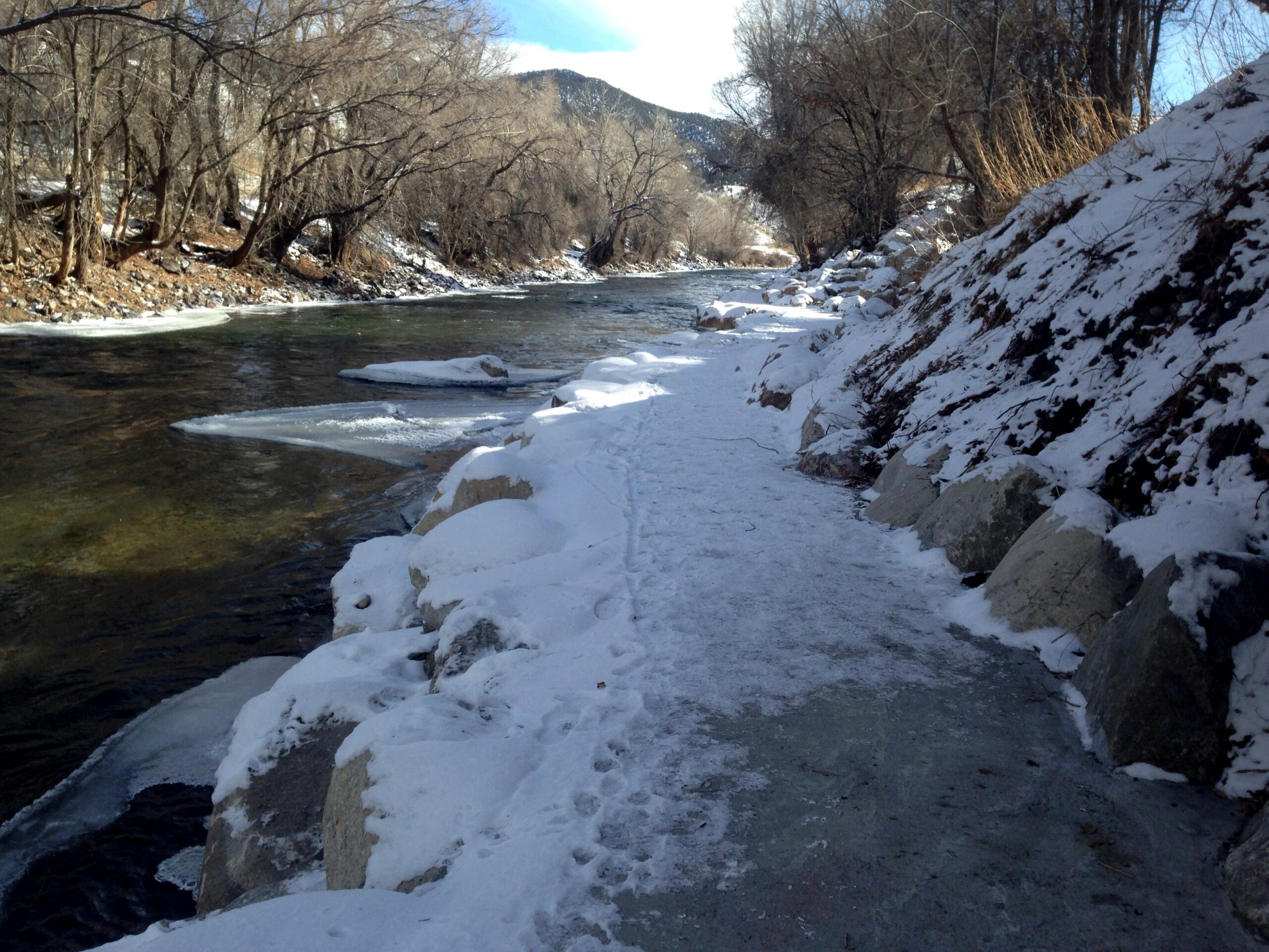 A snowy riverbank with a partially frozen river, lined with bare trees. Snow covers the path and rocks along the water