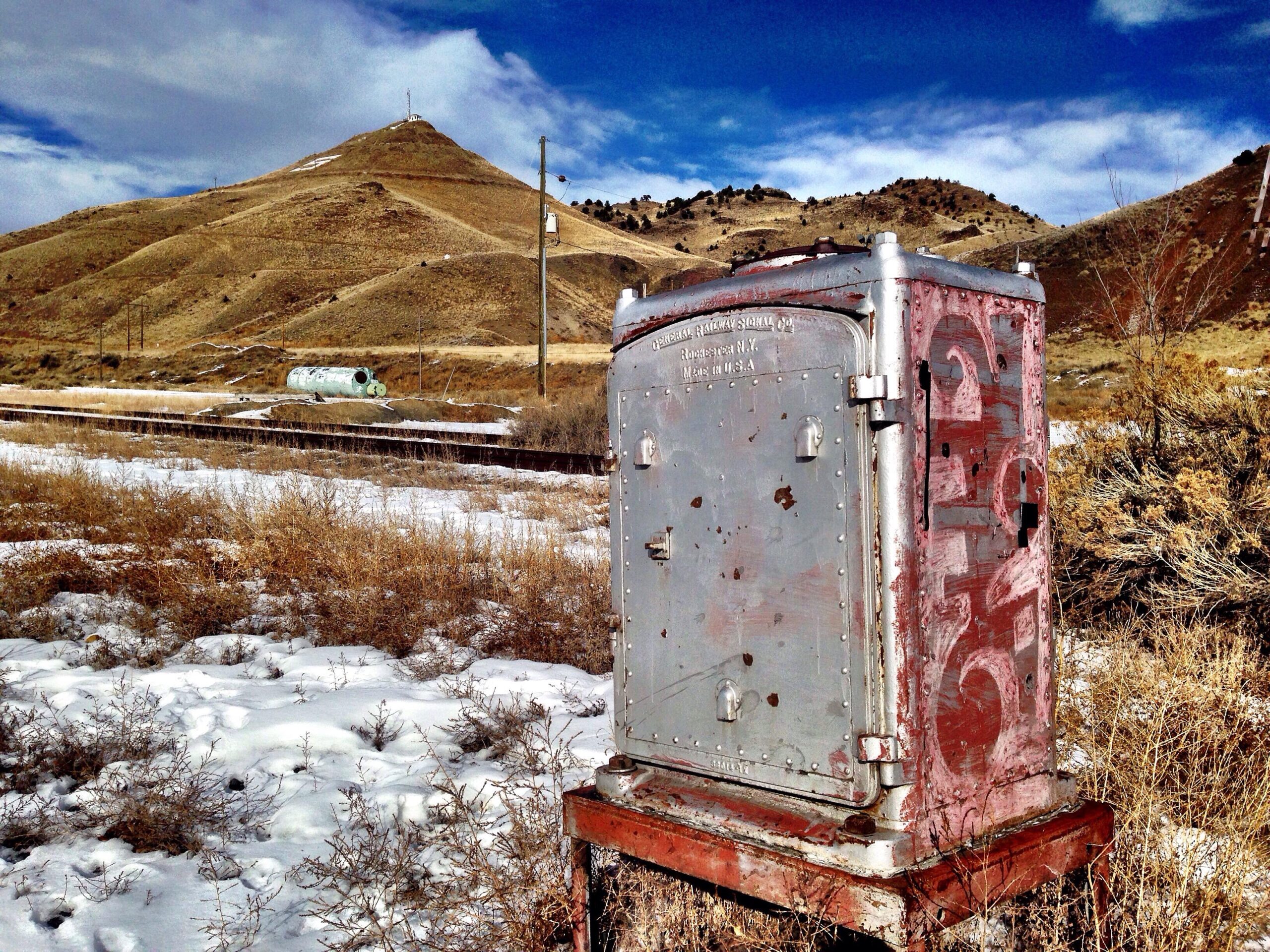 A weathered metal utility box sits in a snowy landscape, with dry grasses surrounding it. In the background, a hillside rises, dotted with sparse vegetation and a few power poles. A green cylindrical object is visible near the railway tracks, with a blue sky partially covered by clouds overhead. Arkansas Hills mountain bike trail.