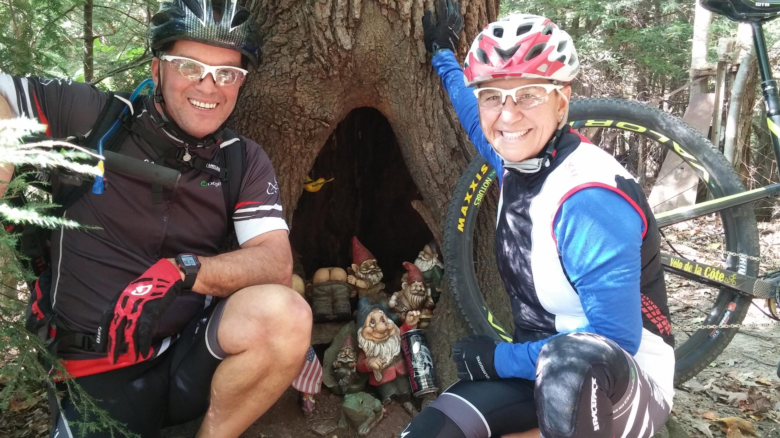 A man and a woman, both wearing cycling gear and helmets, pose next to a large tree with a hollow that contains several colorful garden gnomes. The man is leaning against the tree, smiling, while the woman kneels beside him, also smiling. In the background, a mountain bike is visible. The scene is set in a lush, wooded environment. Mohican mountain bike trail.