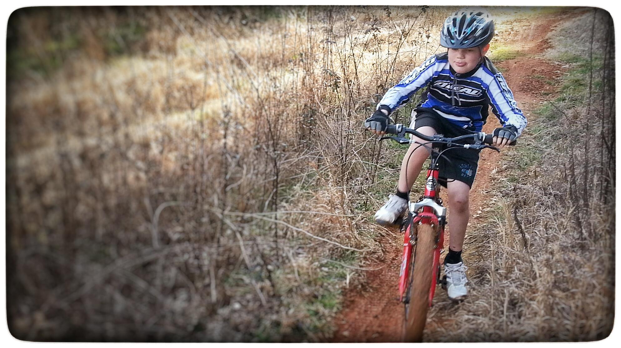 A young boy riding a mountain bike along a dirt path, wearing a helmet and a blue and black jersey. The background features dry grass and sparse vegetation, indicating a natural outdoor setting. Signal Hill mountain bike trail.