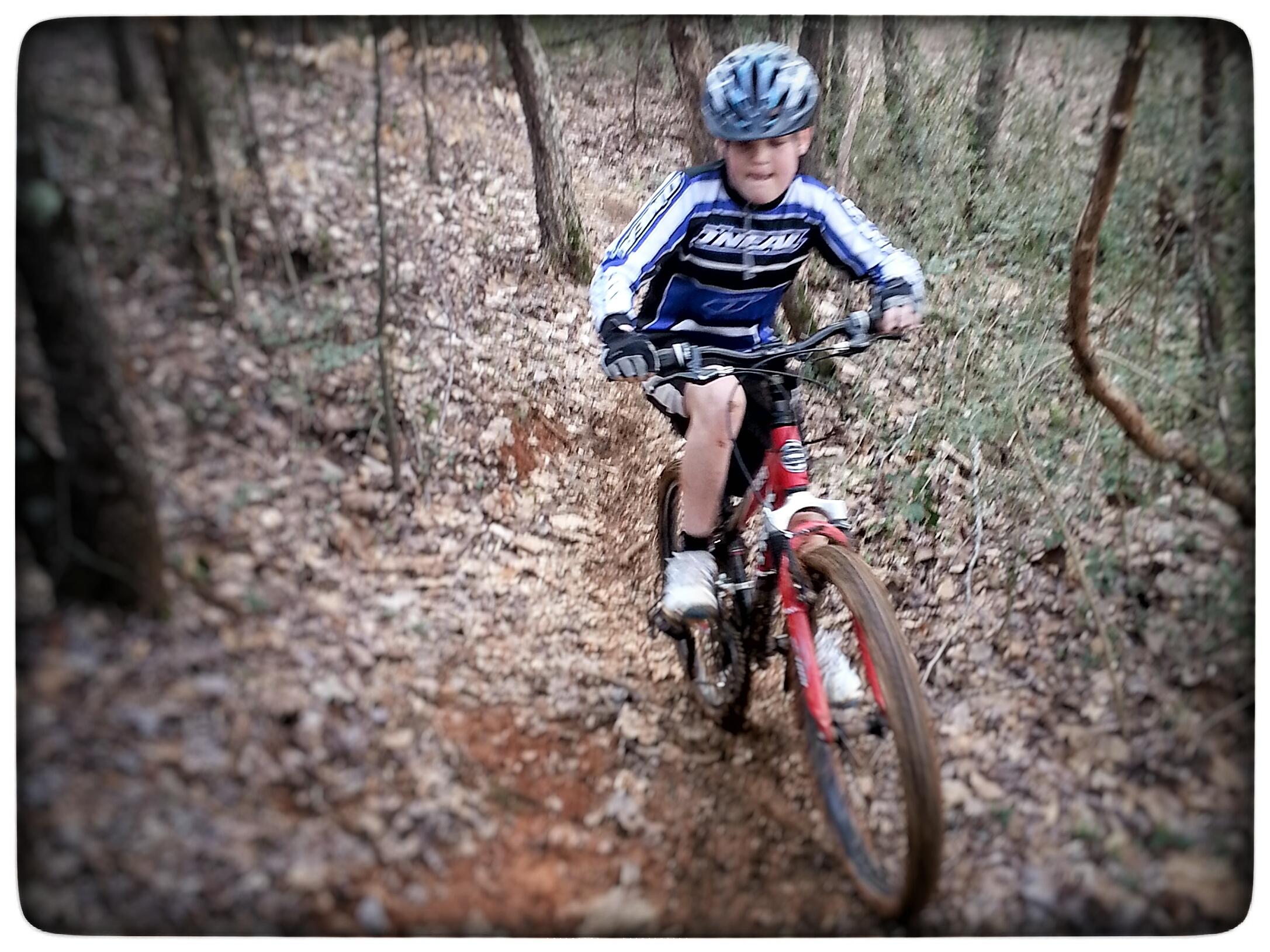 A young boy riding a red mountain bike through a wooded trail covered in leaves. He is wearing a blue and black jersey, shorts, and a helmet, showing concentration as he navigates the rough terrain. The background features trees and underbrush, creating a natural outdoor setting. Signal Hill mountain bike trail.