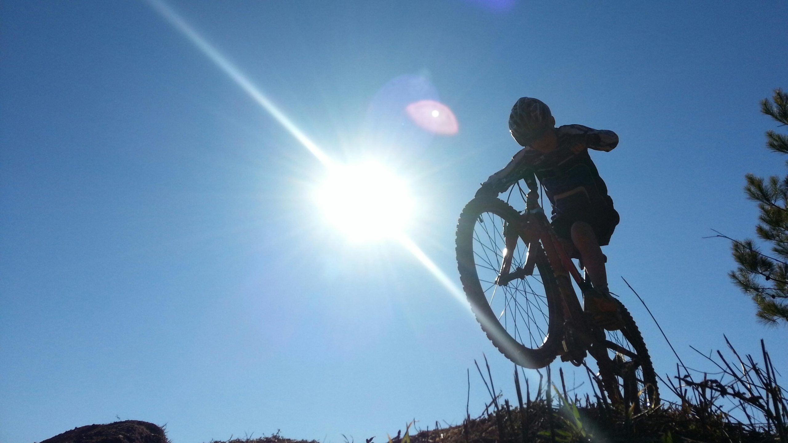 A silhouette of a child performing a bicycle trick against a bright sun in a clear blue sky. The child is balancing on the back wheel of the bike, showcasing a sense of adventure and excitement. Signal Hill mountain bike trail.