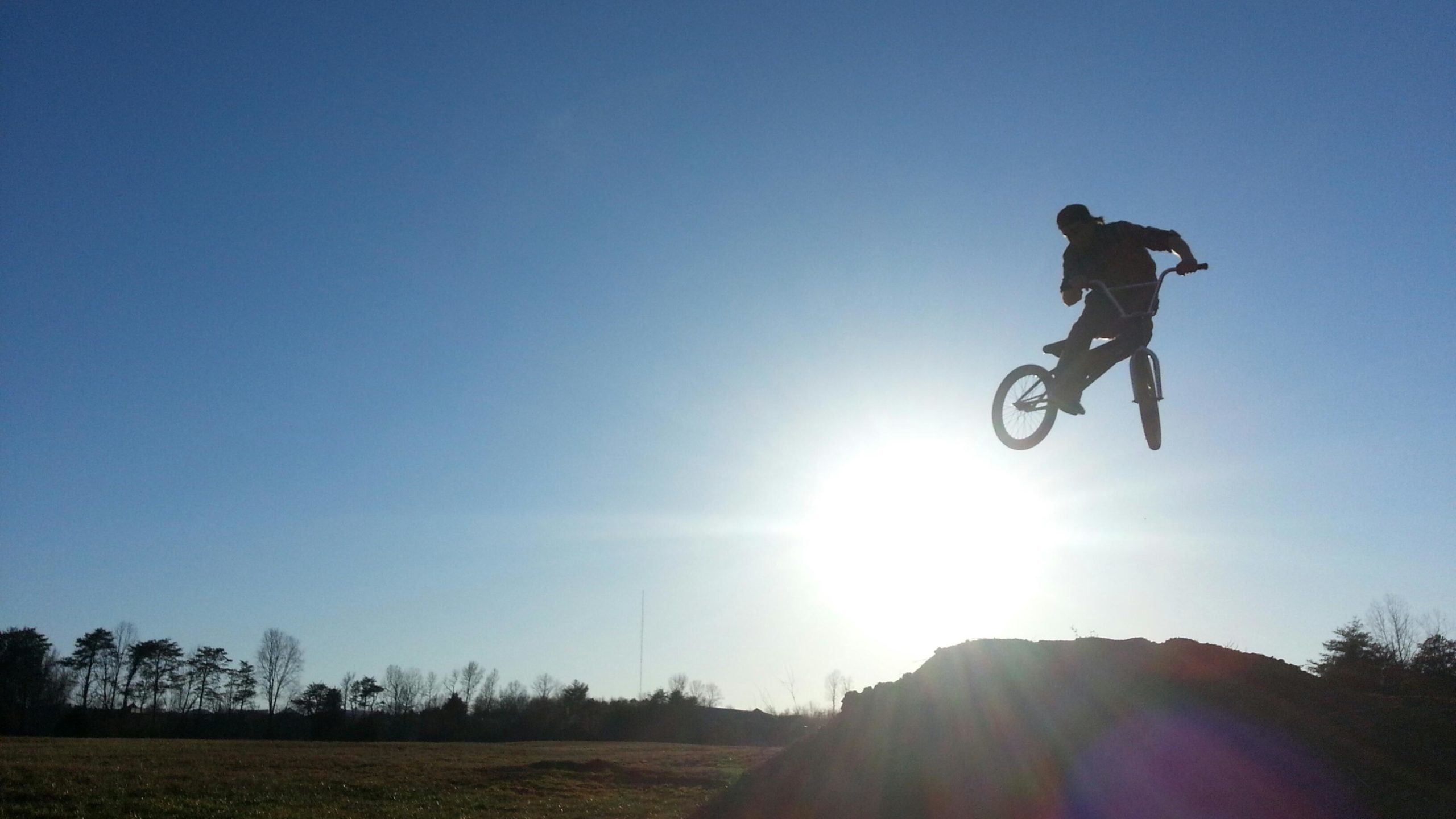 A silhouetted BMX rider performing a jump against a bright sun and clear blue sky, with an open field and trees in the background. Signal Hill mountain bike trail.