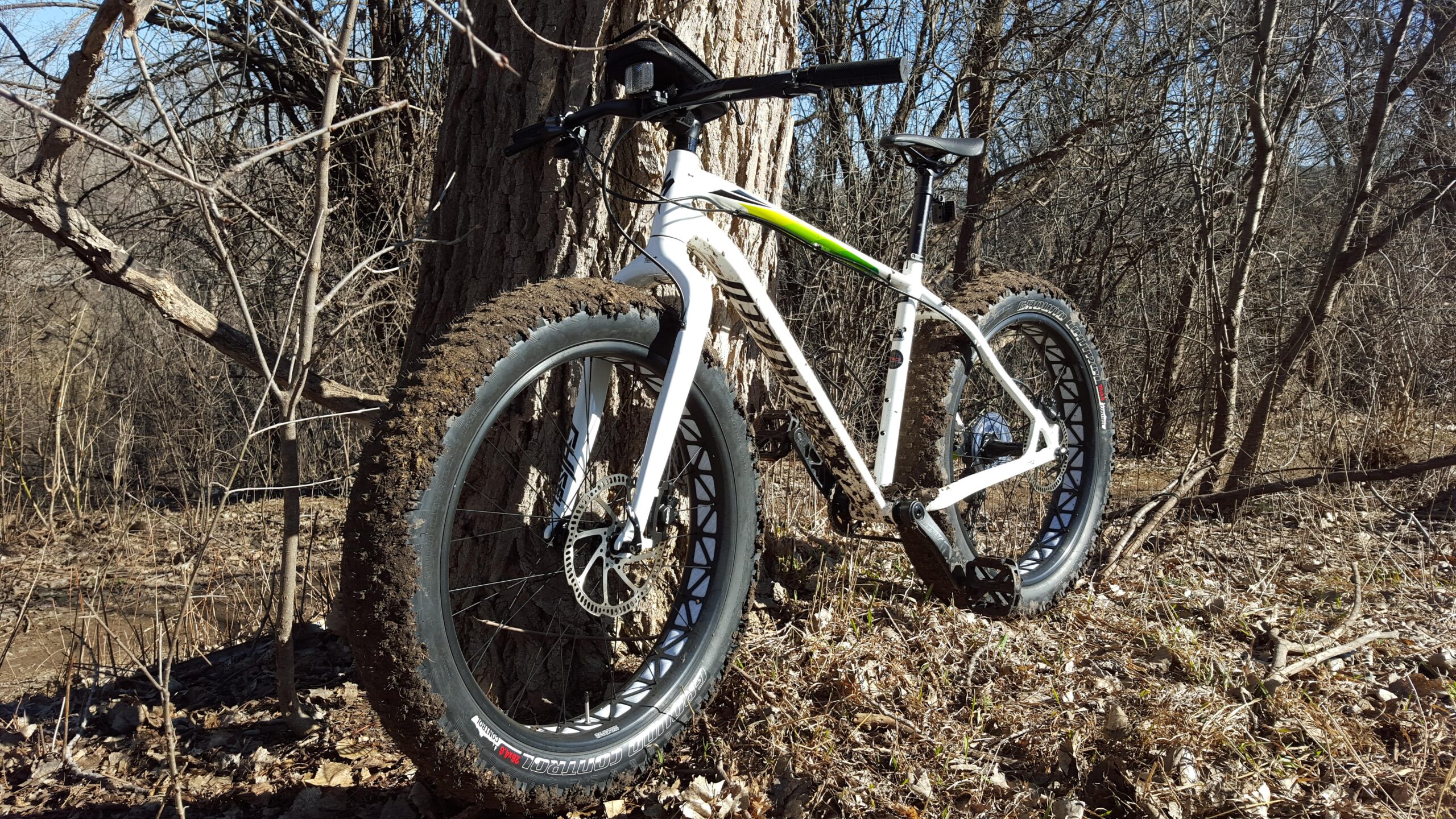Specialized Fatboy: A mountain bike with wide tires leaning against a tree in a wooded area. The bike is partially covered in mud, indicating recent outdoor use. Dry leaves and twigs are scattered on the ground.