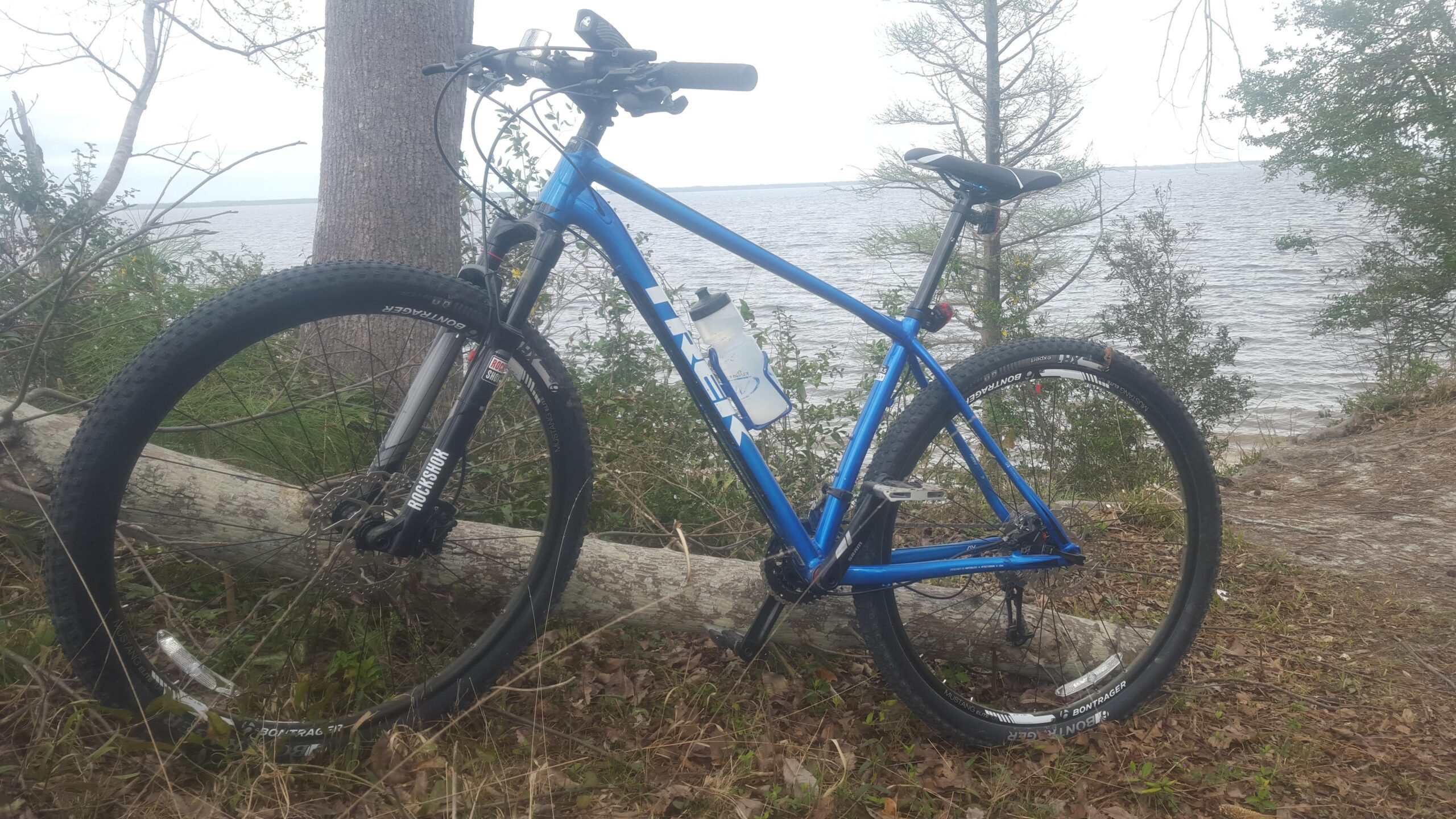 Trek Superfly: A blue mountain bike is resting against a fallen tree near a body of water. The bike features a water bottle attached to the frame and is surrounded by greenery and fallen leaves. In the background, the water stretches out under a cloudy sky.