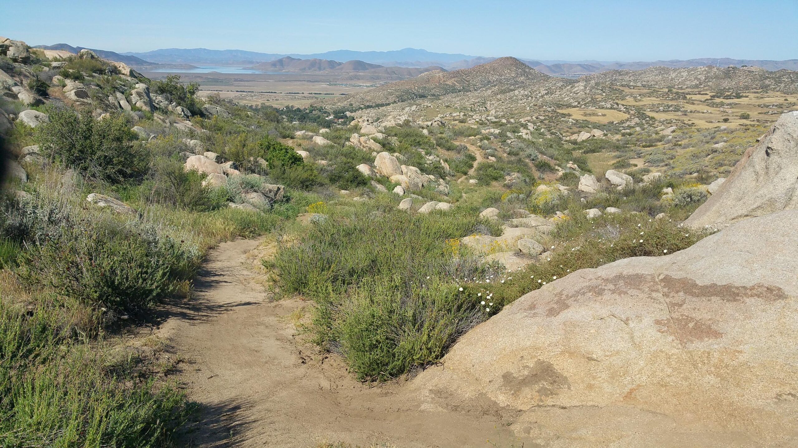 A scenic view of a rocky hillside trail surrounded by green shrubs and wildflowers, leading towards a distant landscape of hills and a body of water under a clear blue sky. Simpson Park mountain bike trail.