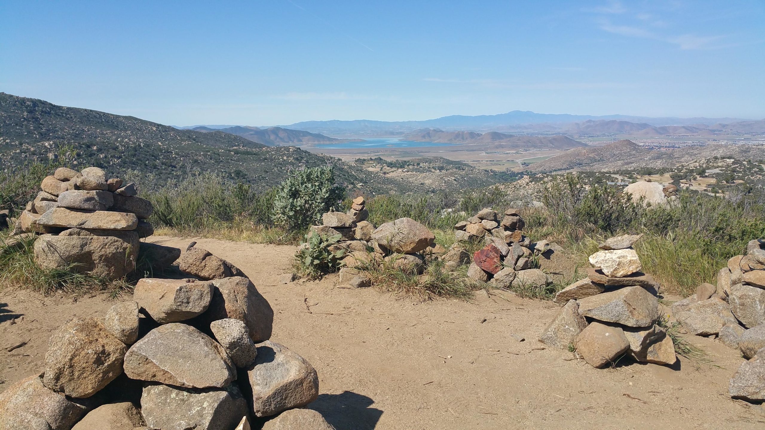 A scenic view from a hiking trail featuring several stacked stone cairns along a sandy path. In the background, rolling hills and a body of water are visible under a clear blue sky. Simpson Park mountain bike trail.